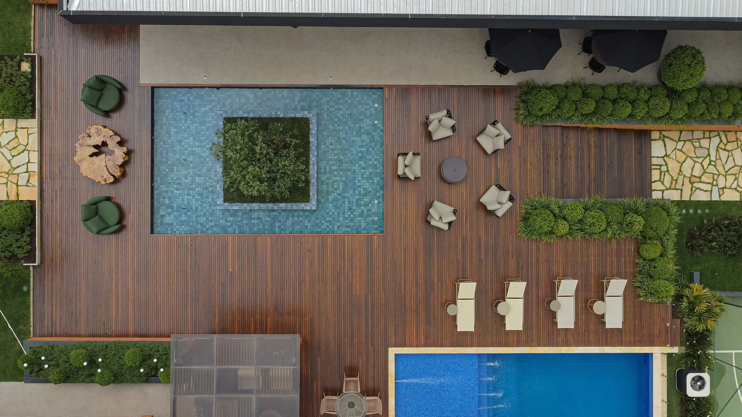 Visão aérea de um deck de madeira com uma piscina em formato de quadrado central, cercada por plantas, cadeiras de descanso, mesas, e guarda-sol.