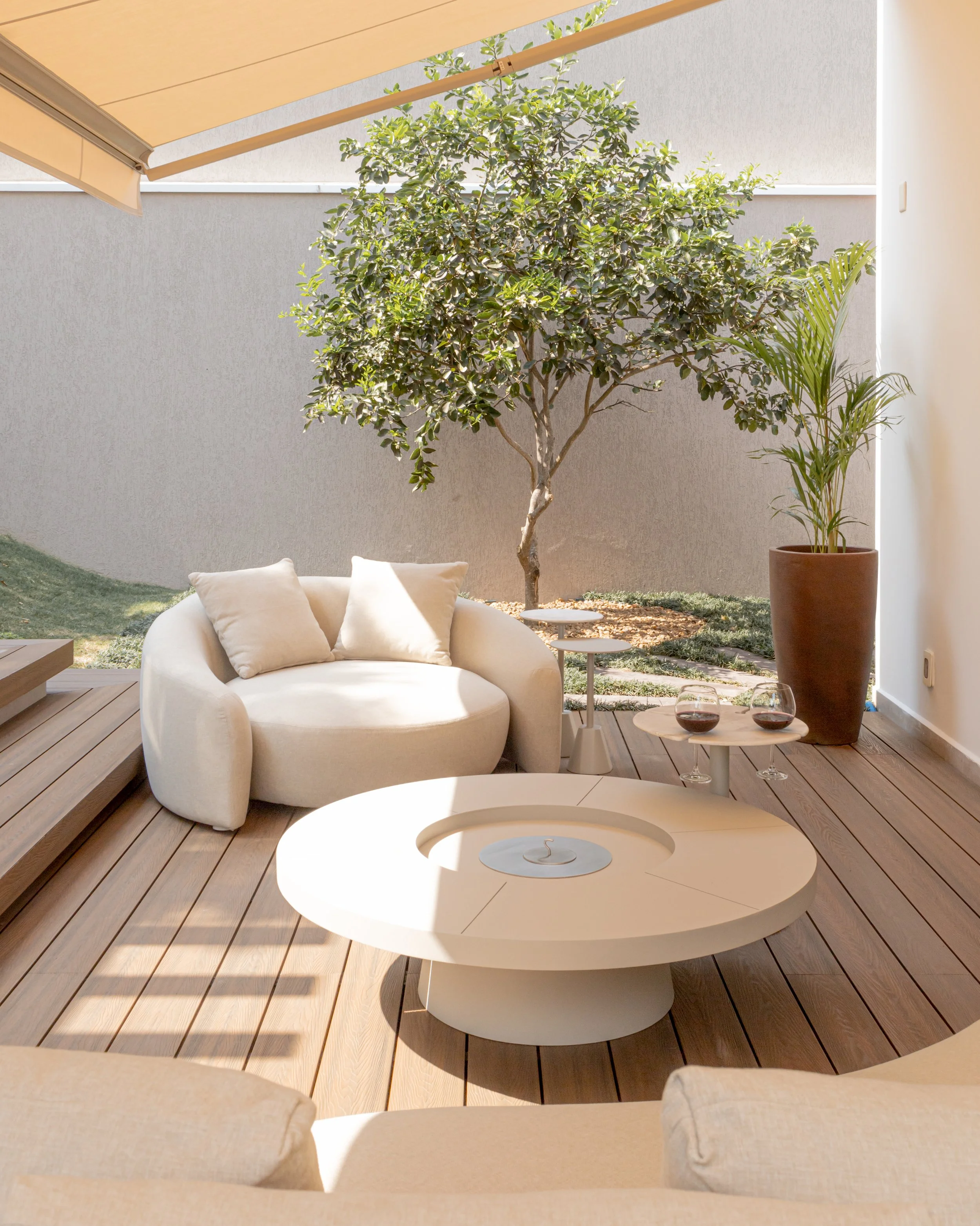 Sala de estar ao ar livre com sofá de almofadas, mesa de centro branca, uma árvore e plantas em vasos, sob um toldo na varanda com piso de madeira.