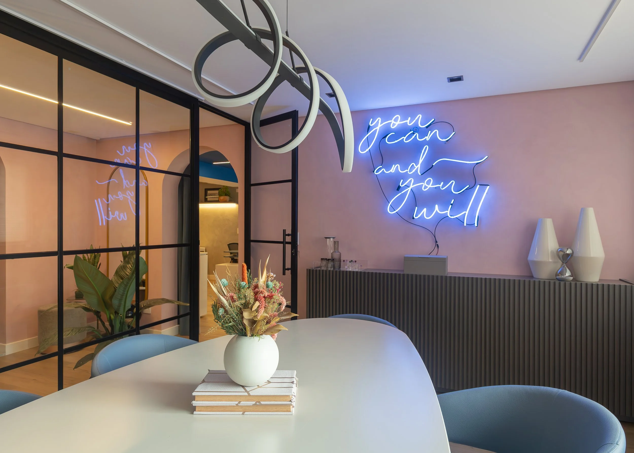 Sala de reunião moderna com mesa branca, vaso de flores coloridas, decoração com iluminação neon azul na parede com frase motivacional, e ambiente decorado com plantas e objetos decorativos.