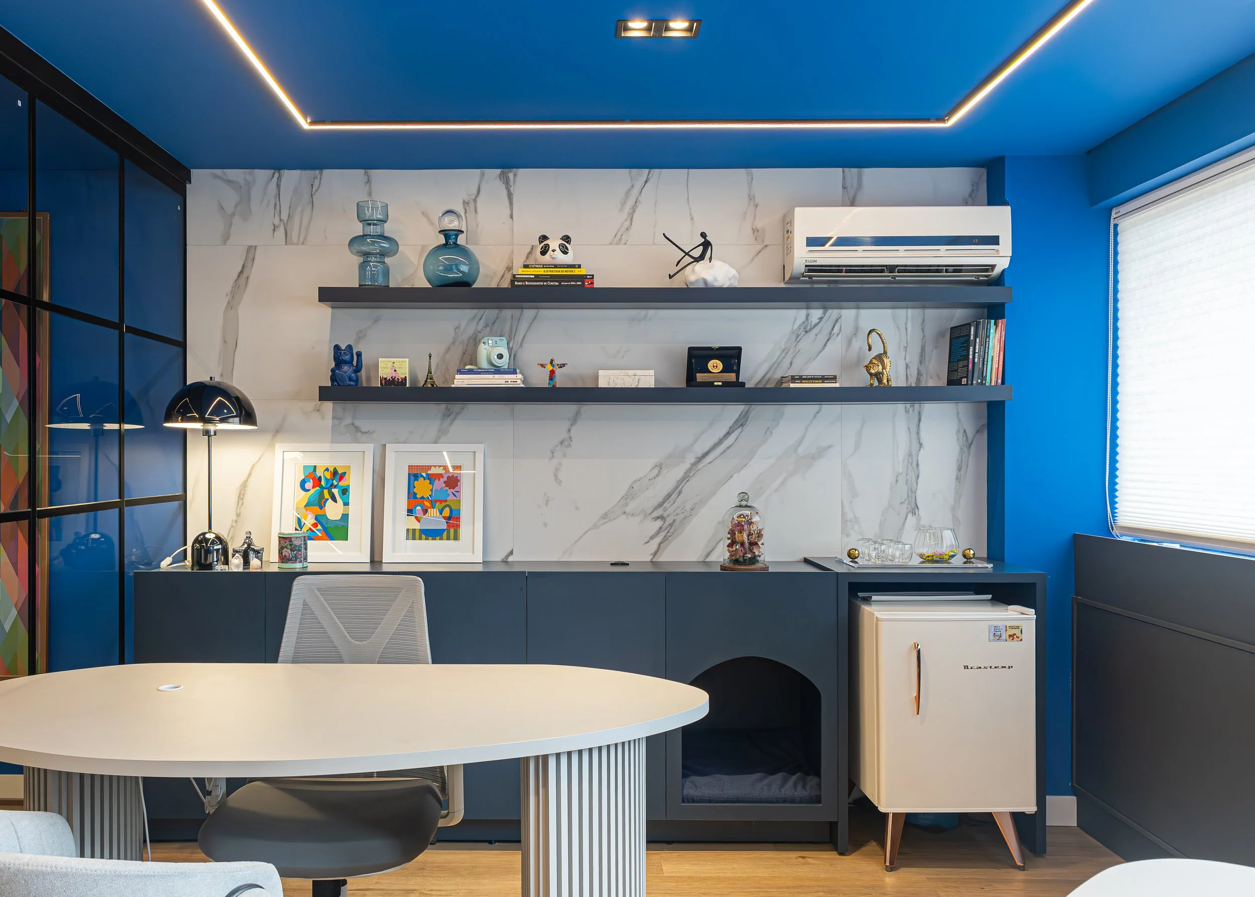 Sala de estar moderna com decoração colorida, prateleiras e objetos decorativos, incluindo vasos e livros, parede de mármore branca, móveis em azul e branco, janela com persiana, e luminária de mesa.