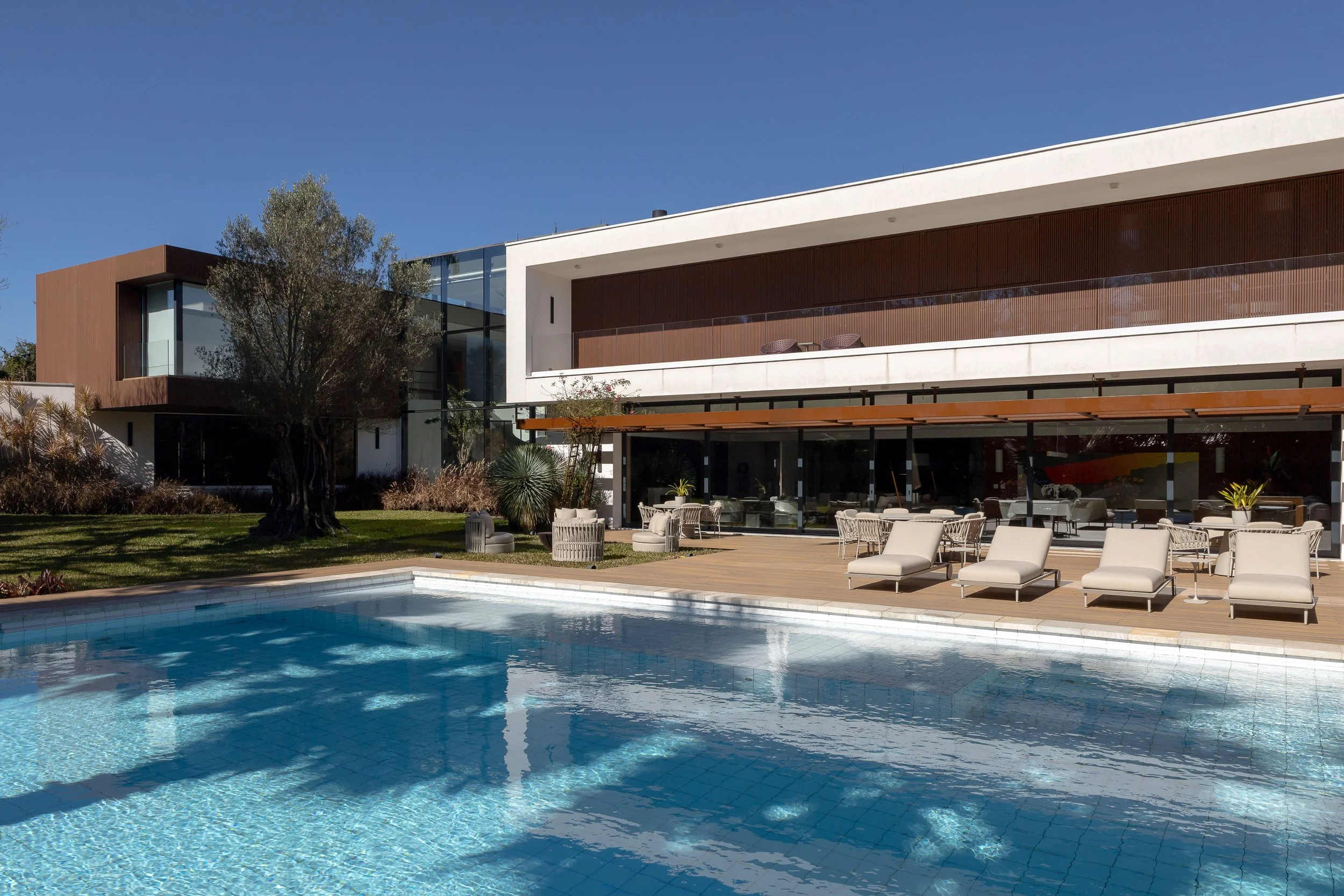 Piscina de frente para uma casa moderna com área de descanso com cadeiras e espreguiçadeiras, jardim com árvores e plantas, céu claro.
