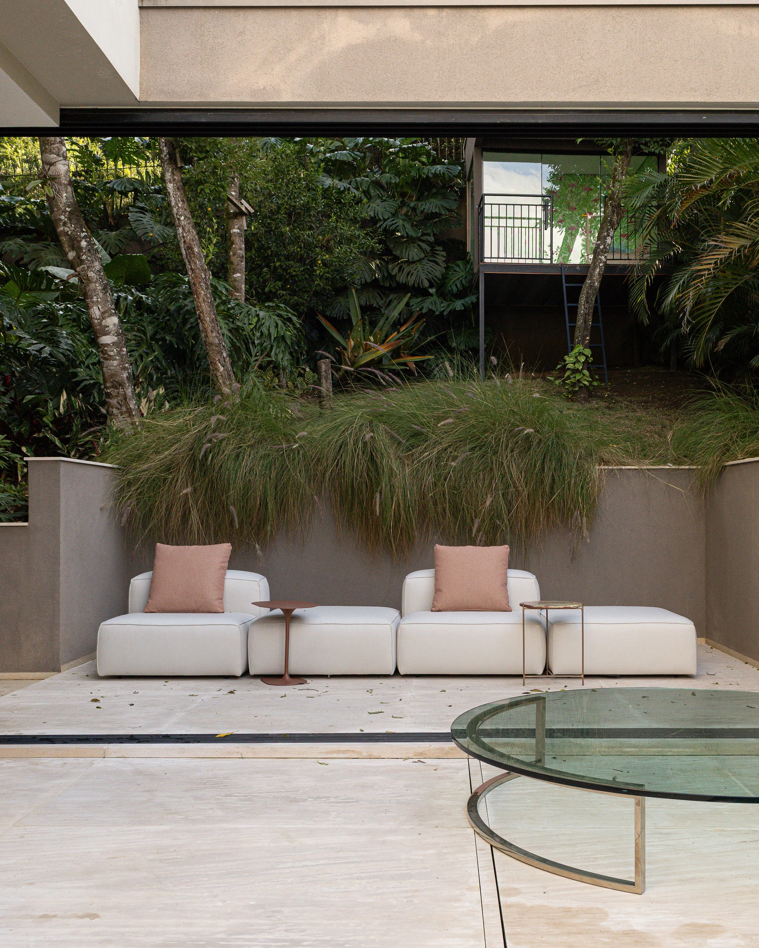 Conjunto de móveis com três assentos brancos, dois com almofadas cor de rosa claro, em um espaço ao ar livre, com vegetação e plantas ao fundo, no interior de uma residência moderna.
