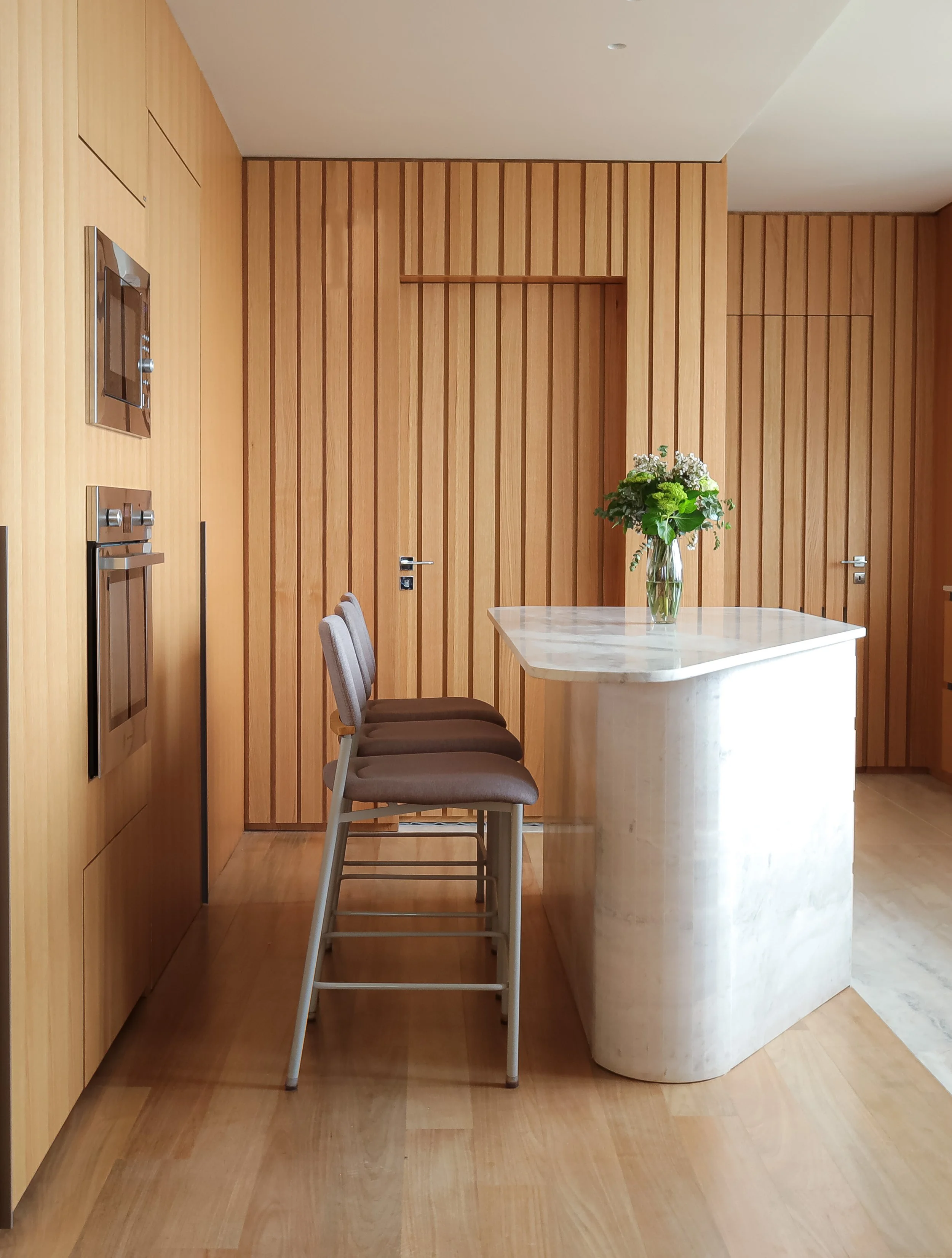Cozinha com paredes de painéis de madeira, bancada de mármore clara, três banquetas cinza com assentos de tecido, e vaso com flores na bancada.