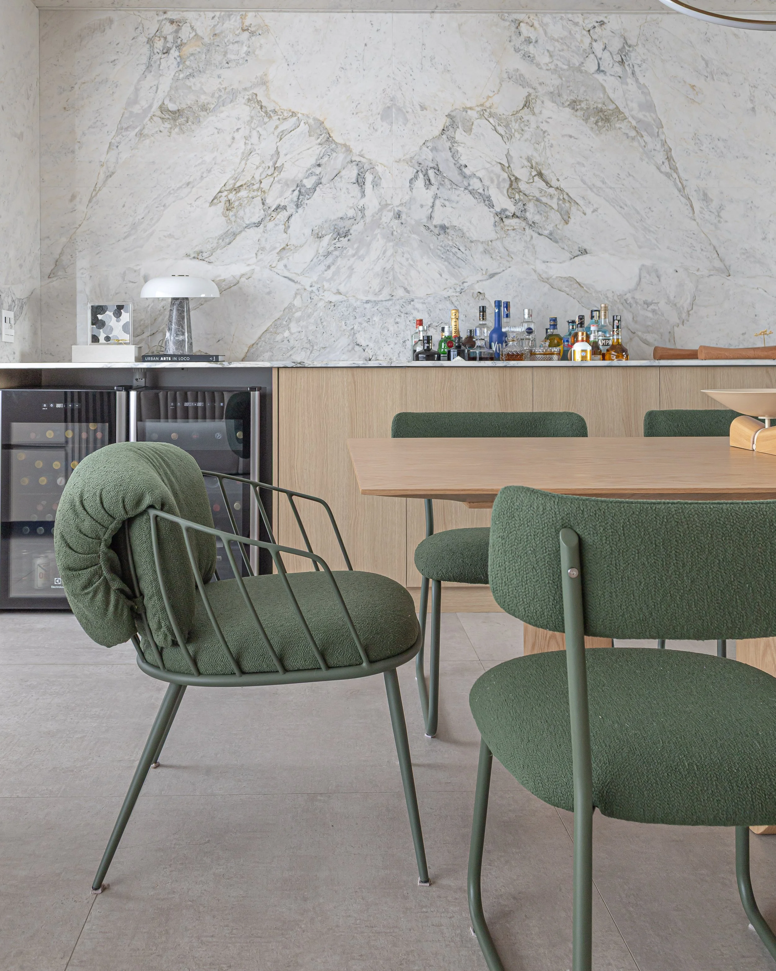 Sala de jantar com mesa de madeira e cadeiras verdes, uma pequena geladeira de vinhos negra, uma bancada com garrafas de bebidas alcoólicas, e uma parede de mármore branco atrás da bancada.