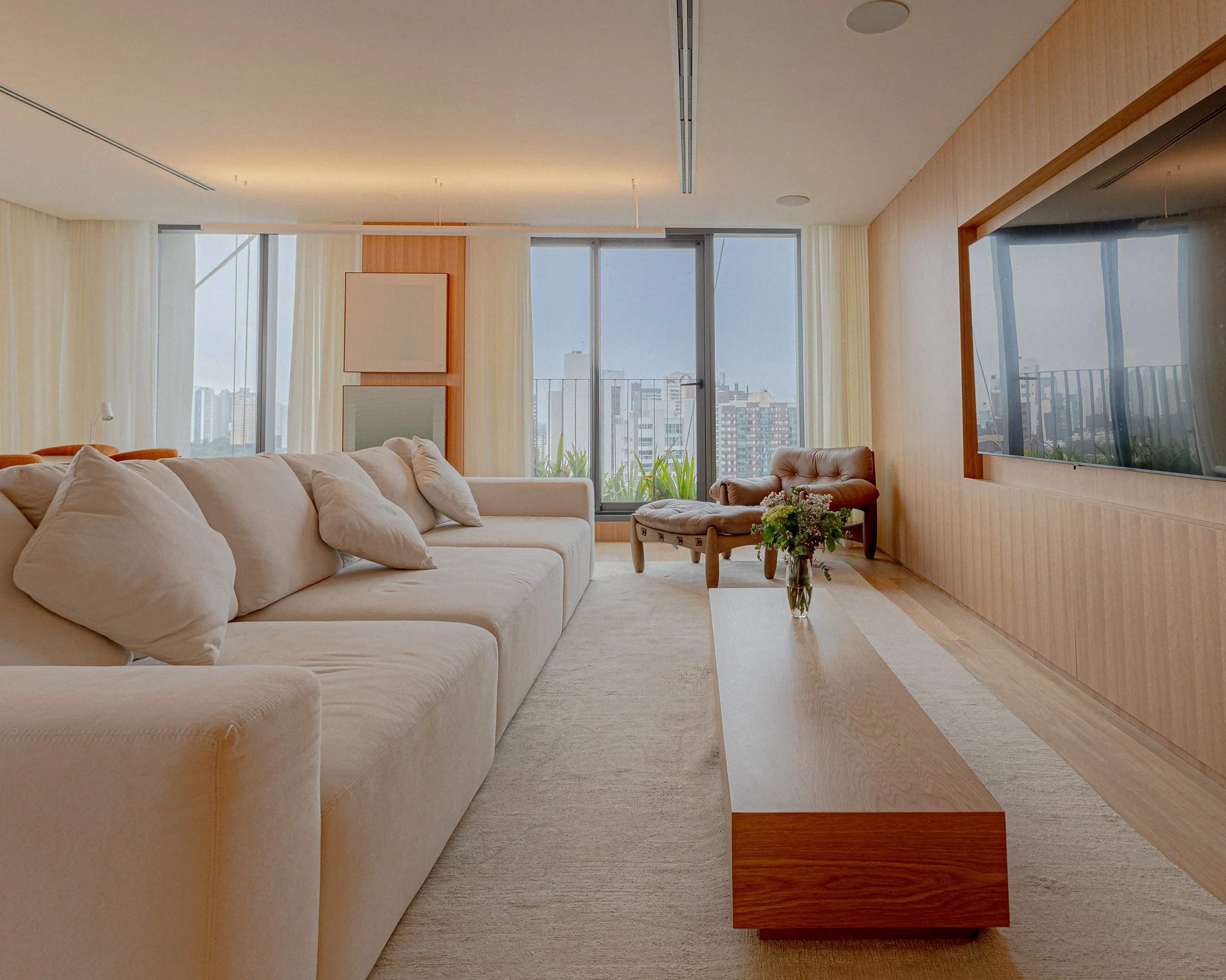 Sala de estar com sofá branco, poltronas de madeira, mesa de centro e televisão de tela grande na parede, com janelas de vidro que revelam edifícios e uma varanda com plantas ao fundo.
