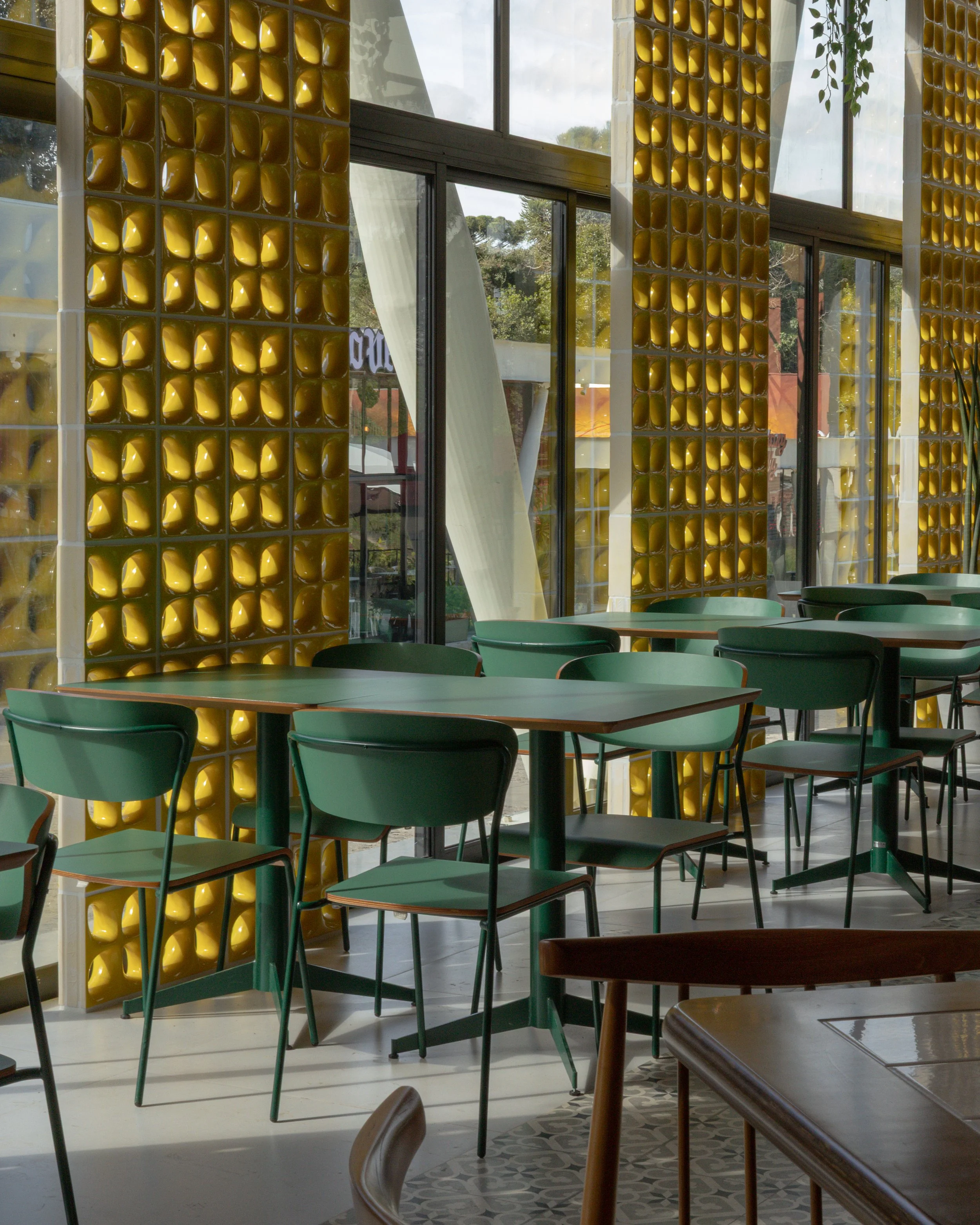 Interior de um restaurante com paredes decoradas com blocos de vidro amarelos e janelas grandes, com várias mesas e cadeiras verdes.