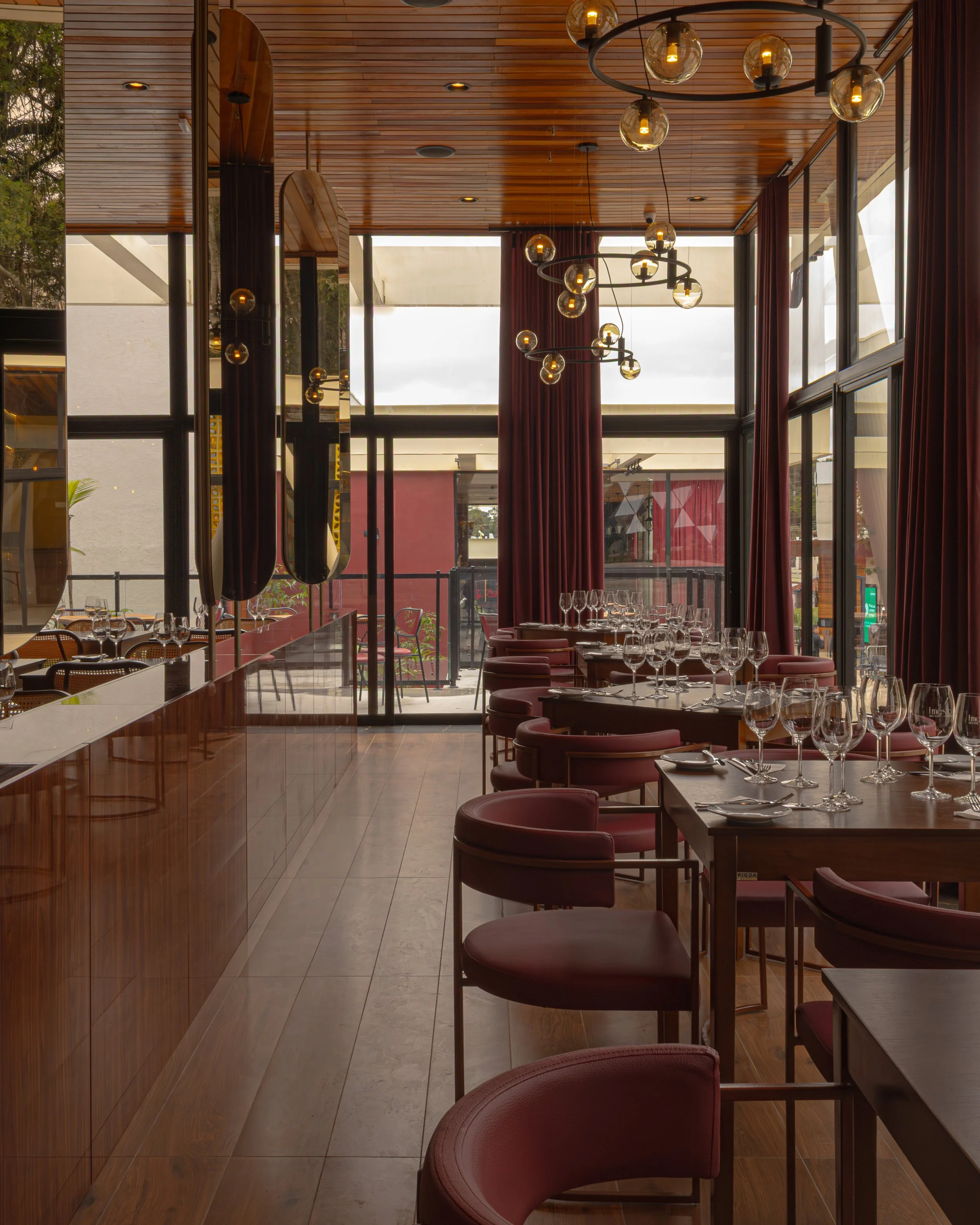 Interior de restaurante com mesas e cadeiras de posti no estilo contemporâneo, cortinas vermelhas, iluminação moderna com lustres de bulbos expostos, paredes de vidro e decoração em tons quentes.