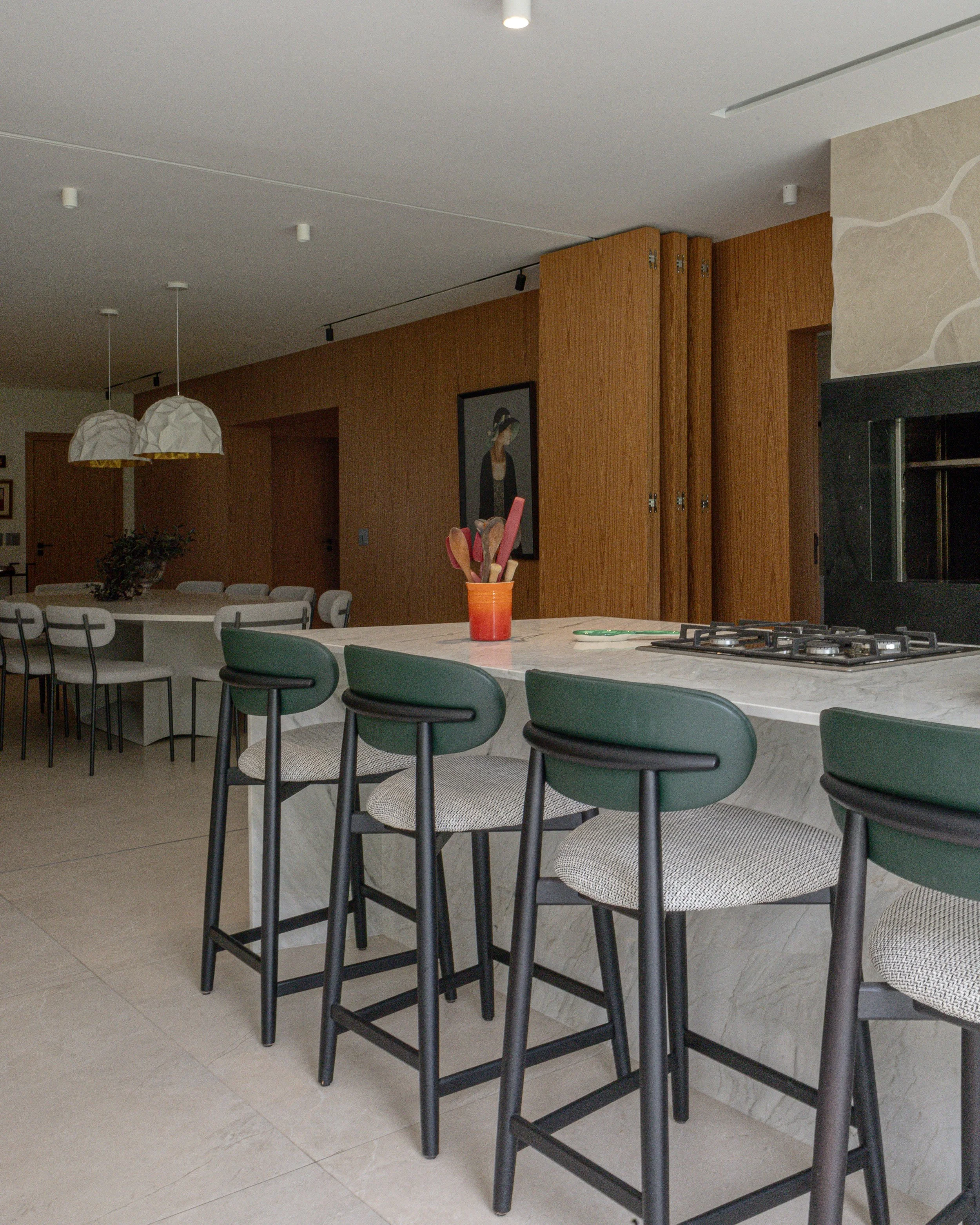 Cozinha moderna com ilha de mármore, cadeiras verdes, lâmpadas pendentes brancas, parede de madeiramento e uma obra de arte na parede.