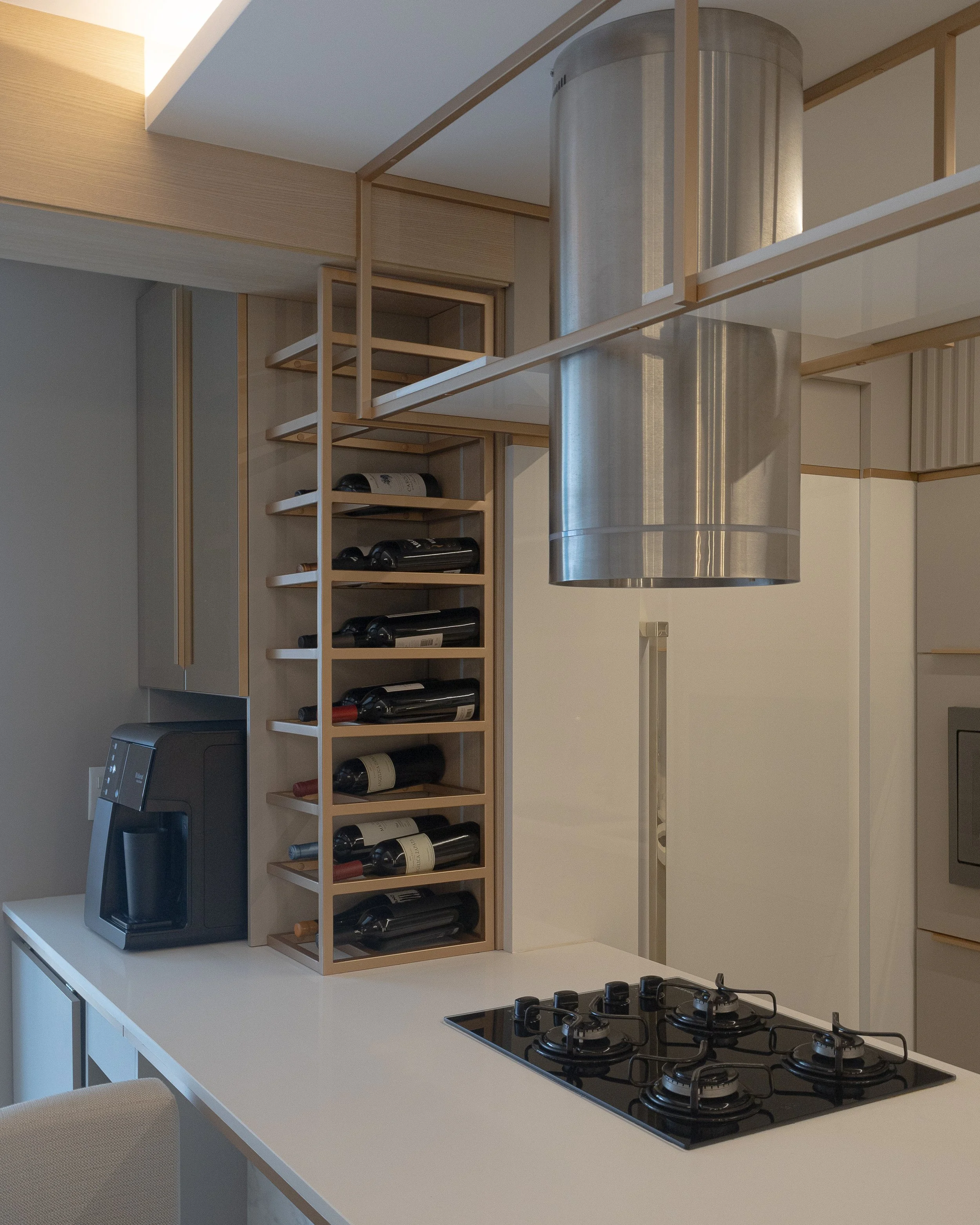 Cozinha moderna com balcão branco, com uma pia de quatro bocas, uma chaleira elétrica e uma torre de armazenamento de vinhos feita de madeira. No teto, um exaustor de metal