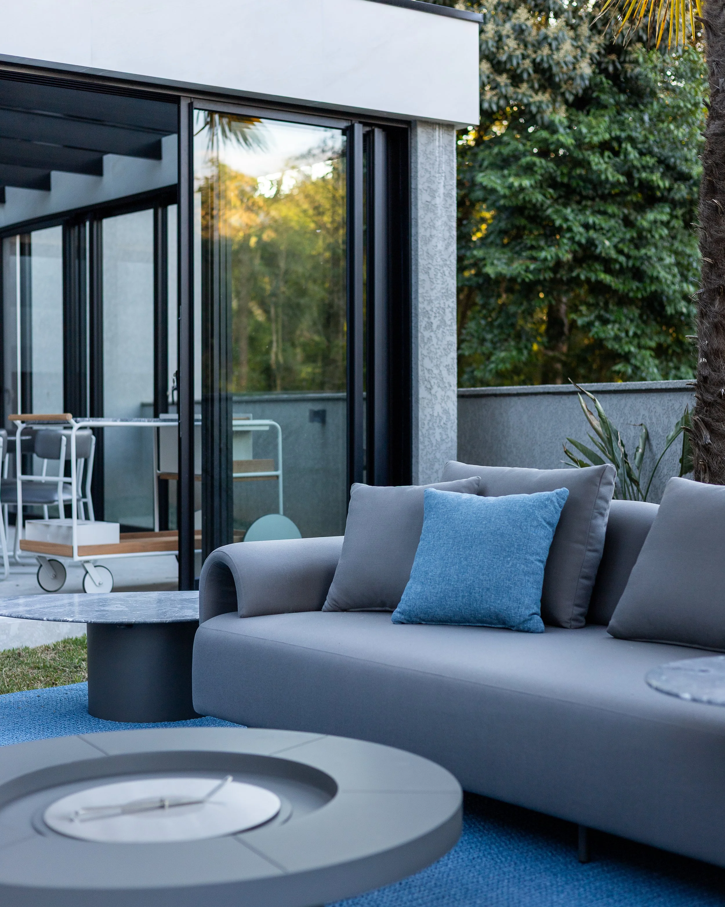 Sacada com móveis de estar, incluindo um sofá cinza com travesseiros azul e cinza, mesa de centro e uma área externa com plantas, árvores e fachada moderna do prédio