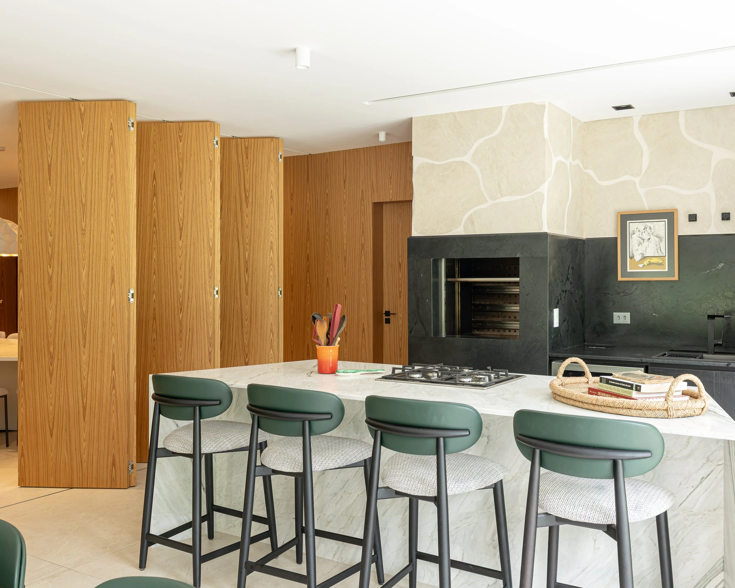 Cozinha moderna com ilha central de mármore, quatro banquetas verdes, paredes de madeira, forno de pizza embutido, quadro na parede e utensílios de cozinha.