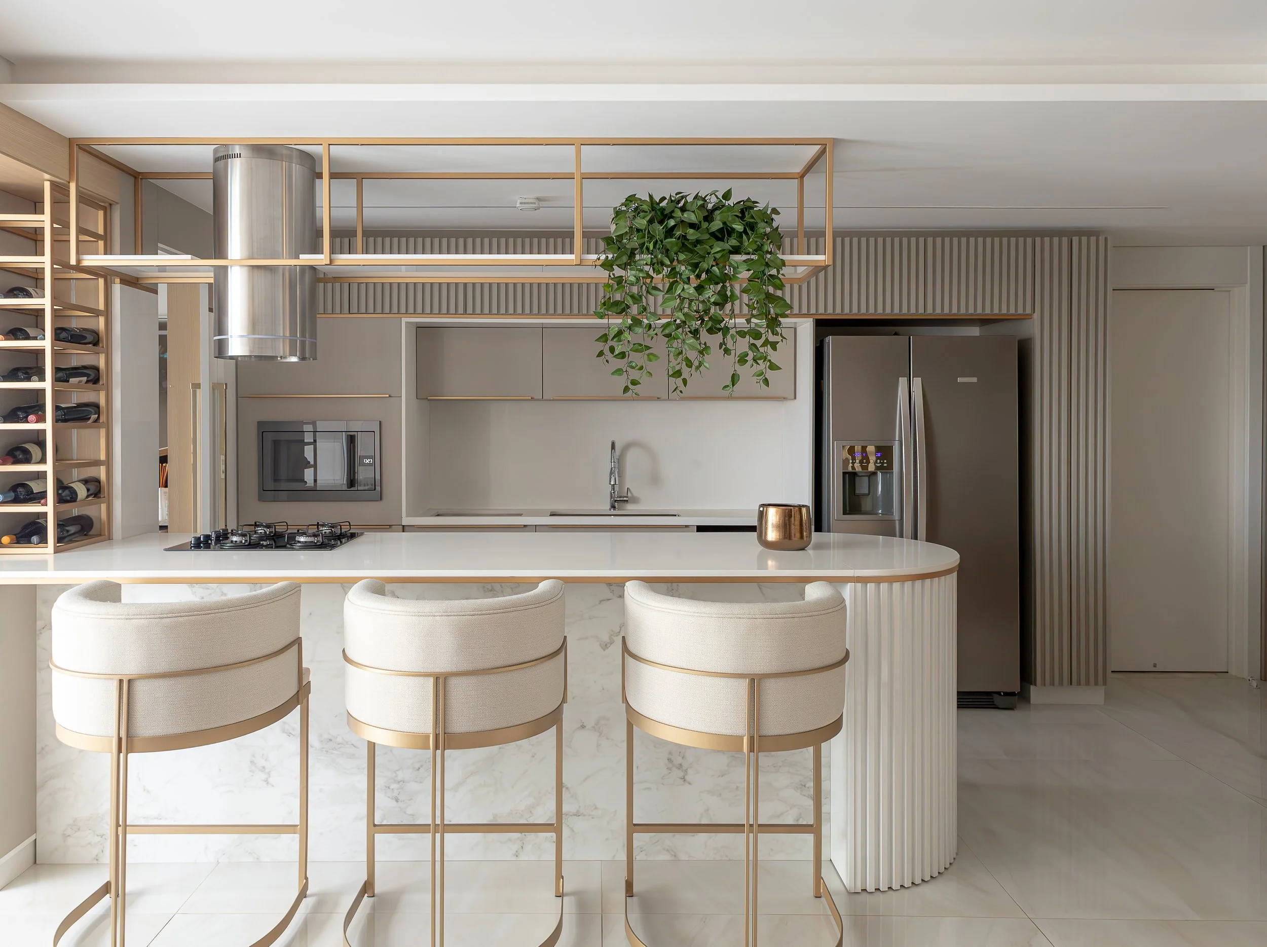 Cozinha moderna com ilha de mármore, três banquetas brancas com detalhes dourados, geladeira de aço inox, armários brancos, prateleiras de madeira, um vaso dourado, planta pendurada e micro-ondas embutido.