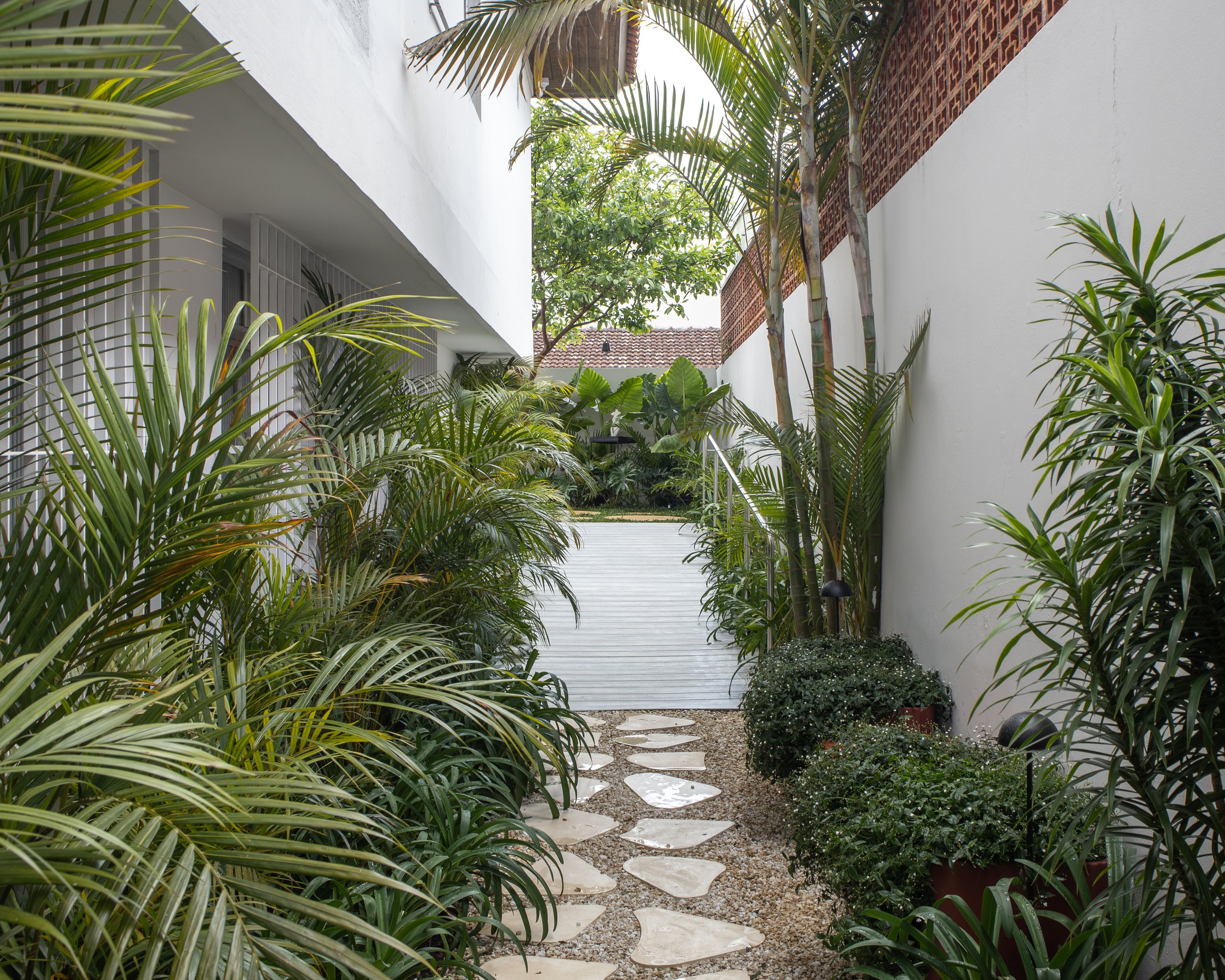 Caminho de pedras na passagem entre plantas verdes e arbustos ao lado de uma parede branca, com árvores ao fundo e uma rampa de acesso.