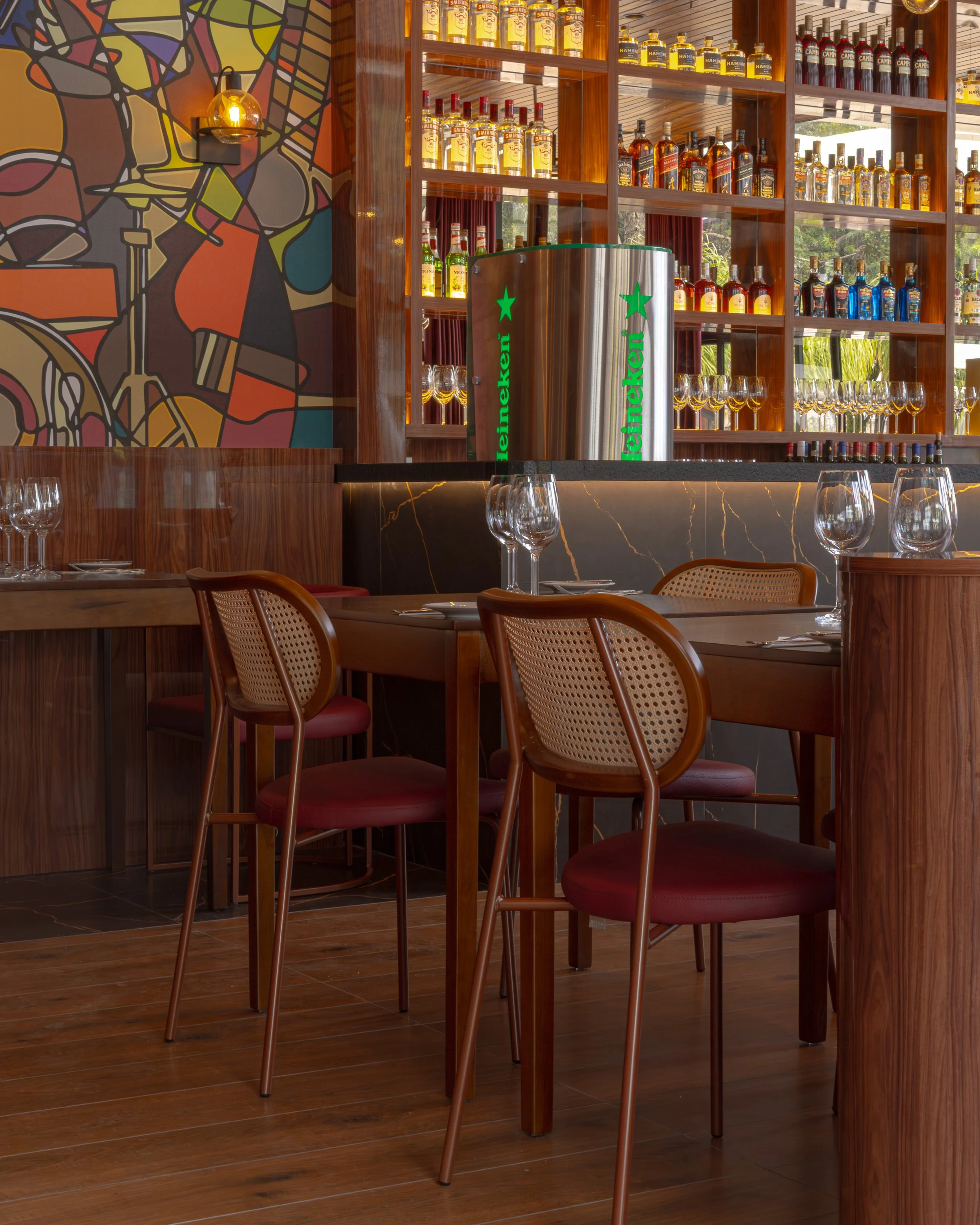 Interior de um restaurante com mesas de madeira, cadeiras de couro vermelho e uma parede decorada com uma arte colorida. No balcão, uma garrafa de Heineken iluminada. Há prateleiras com várias garrafas de bebidas e copos de vinho ao fundo.