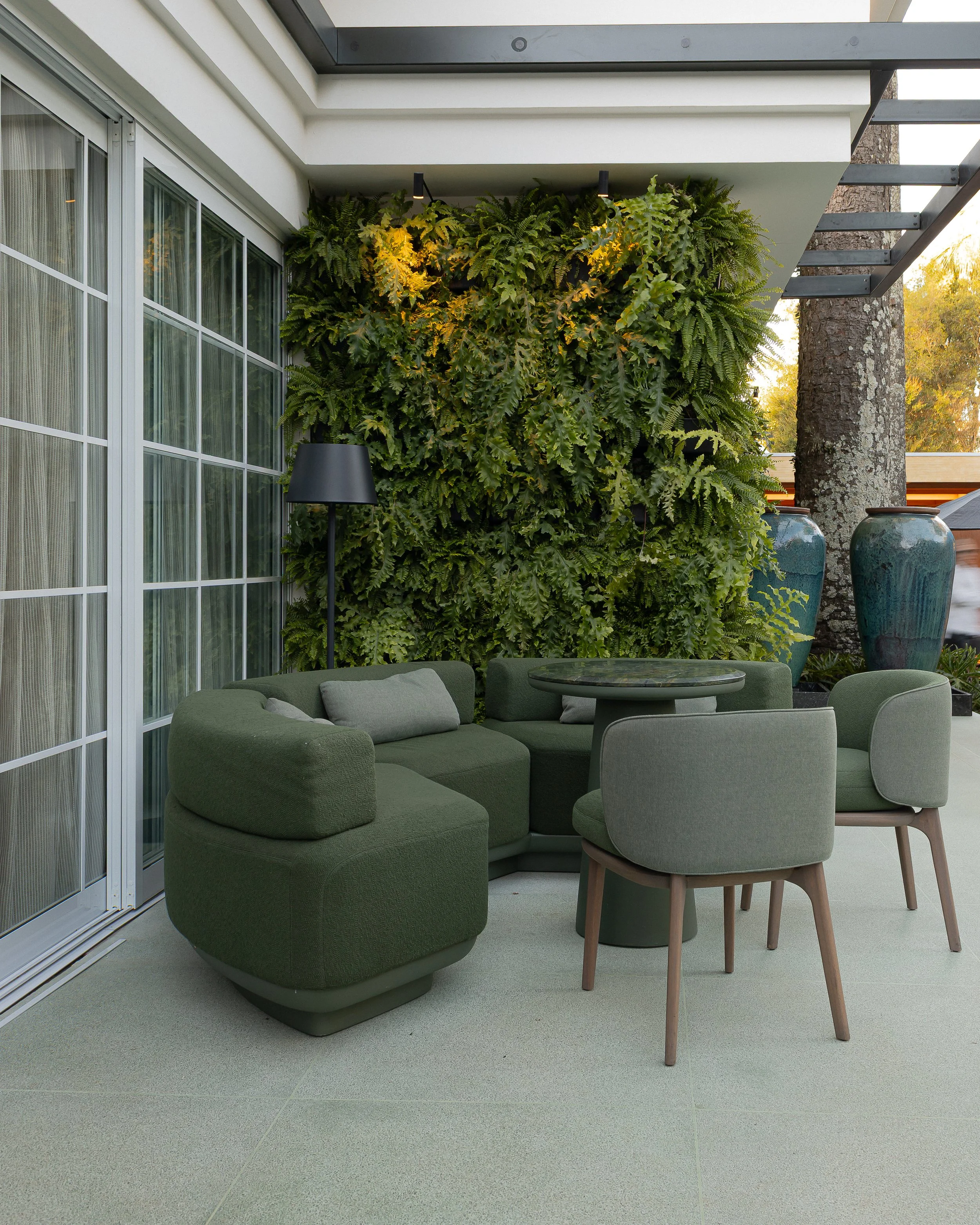 Espaço de convivência ao ar livre com móveis verdes, incluindo um sofá, duas poltronas e uma mesa redonda, com parede de plantas ao fundo e vasos decorativos ao lado.
