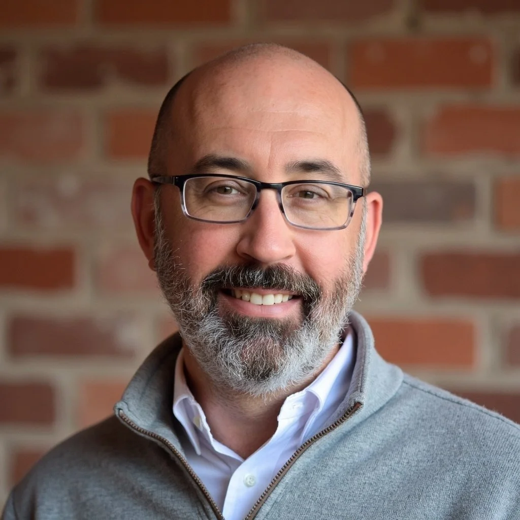 A smiling middle-aged man with glasses, a beard, and a bald head, wearing a white shirt and a gray zip-up sweater, standing in front of a brick wall.