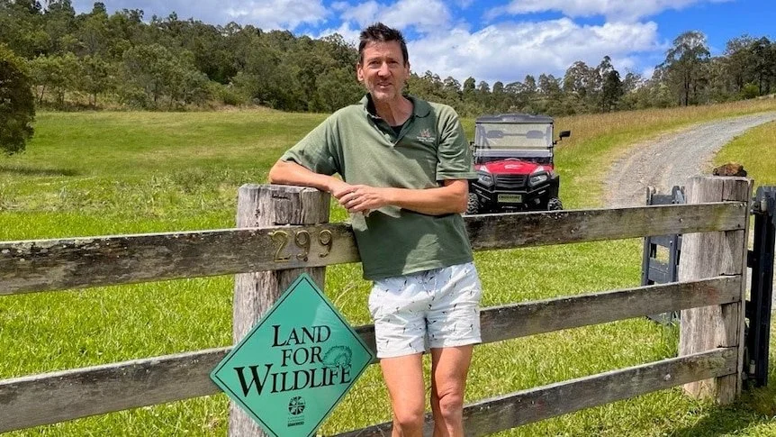 Graham Creed in a green polo shirt and white shorts standing beside a wooden gate with a 'Land for Wildlife' sign, in a grassy field with a golf cart and trees in the background.