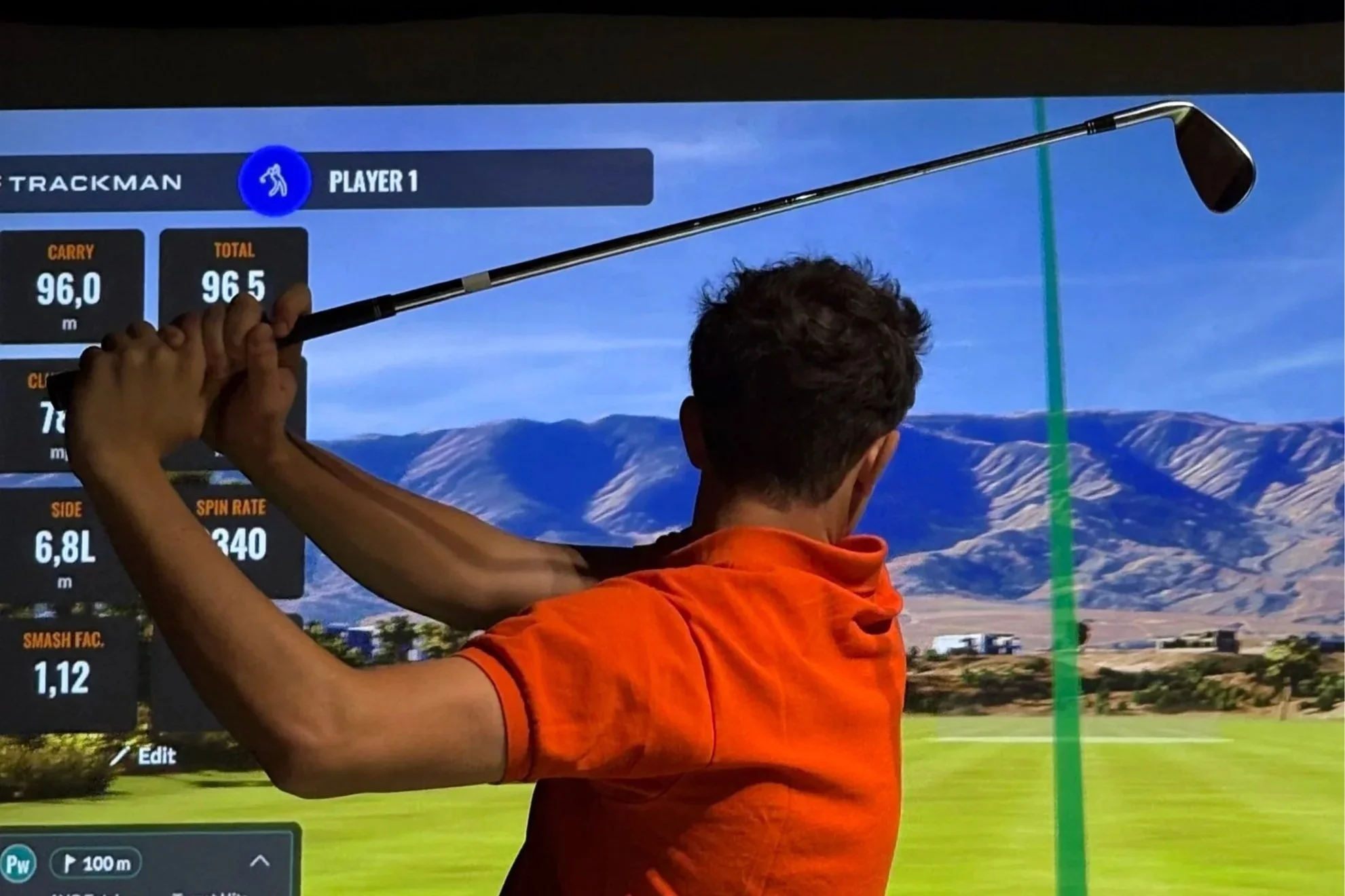 A person dressed in orange holding a golf club and swinging at a golf simulator screen displaying a virtual golf course and player statistics.