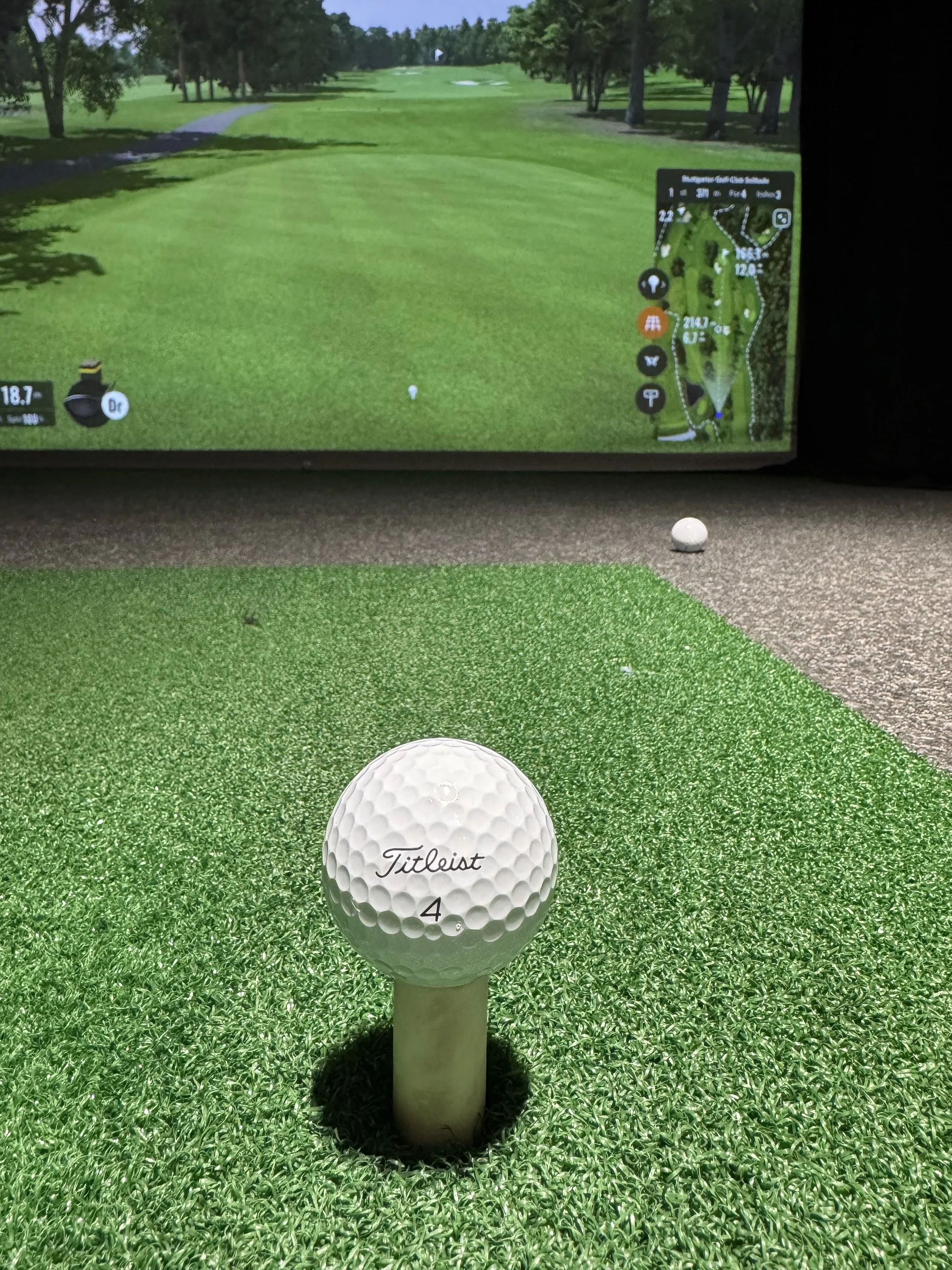 Golf ball placed on a tee on a green artificial turf surface at a driving range, with a digital golf course map displayed on a screen in the background.