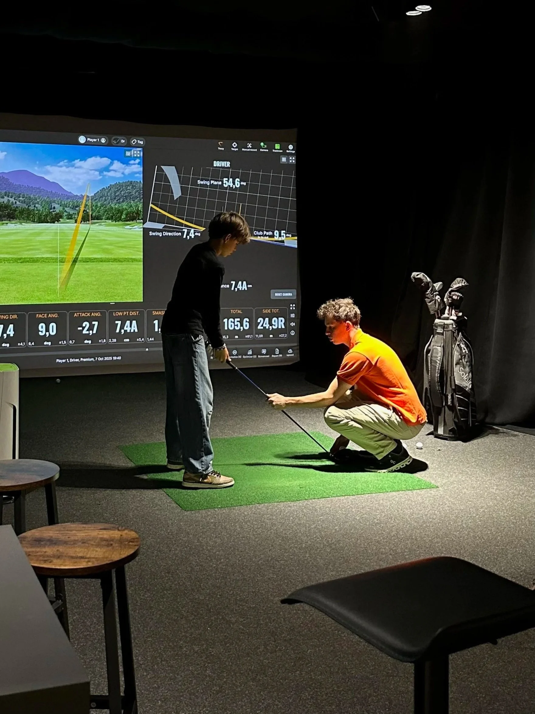 Two individuals in a golf simulator room, one standing and one crouching on a small green mat. The standing person is holding a golf club, while the crouching person appears to be instructing or checking positioning. A large screen displays golf swing data and a simulated golf course in the background, with a golf bag nearby.