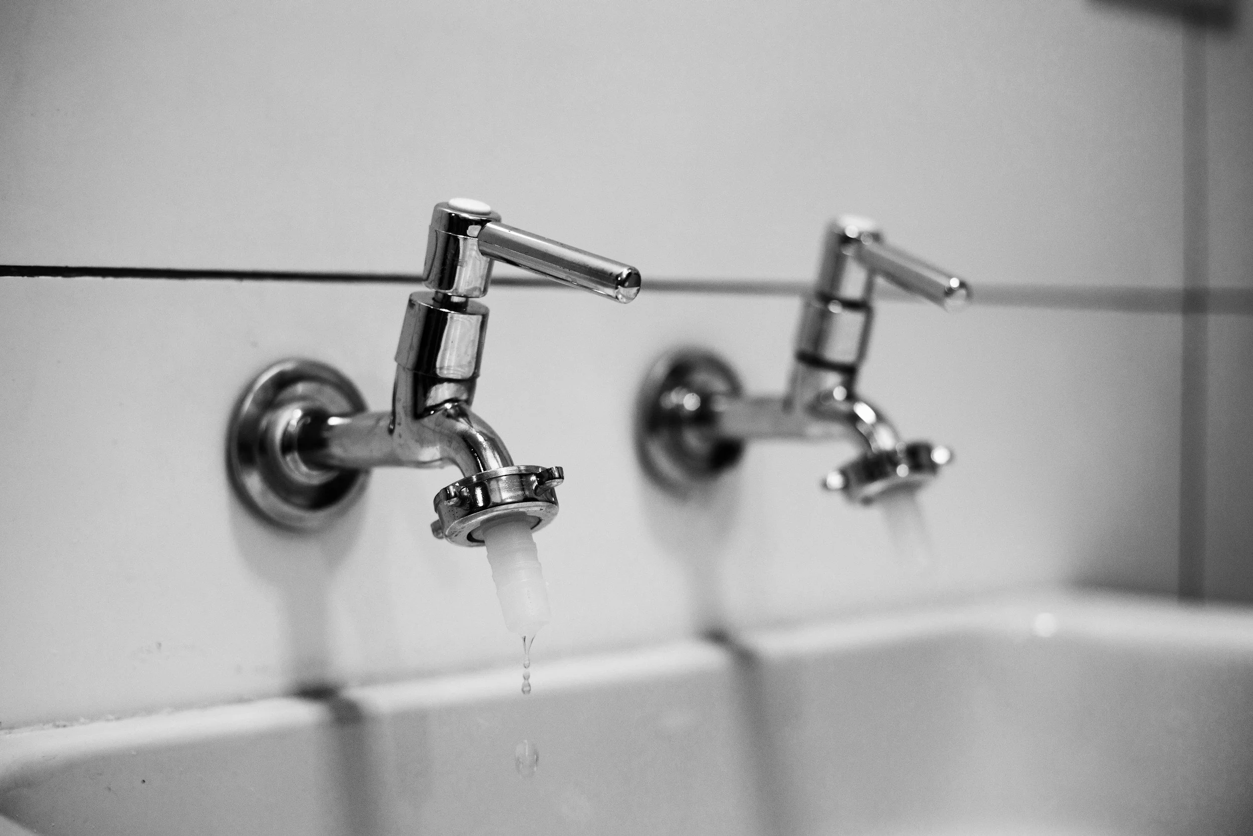 A black and white photo of two metal faucets with water dripping from the spout of the left faucet, mounted on a white tiled wall.