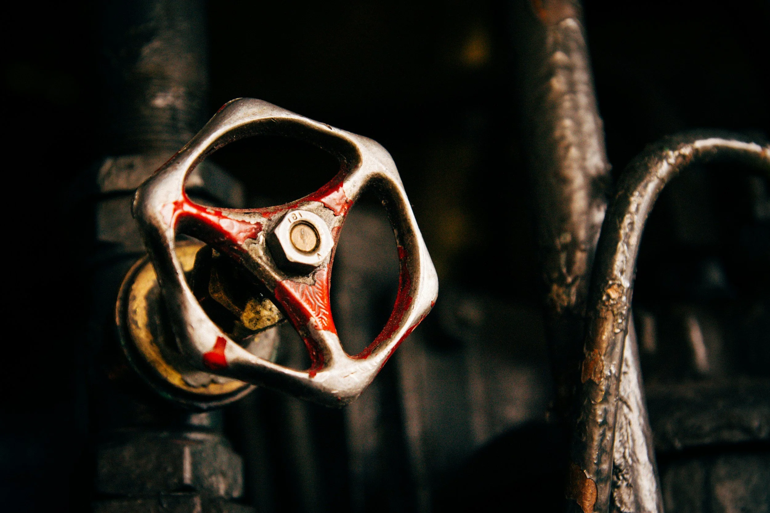 Close-up of a rusty industrial valve or handle with red and silver paint, attached to a dark pipe or machinery.