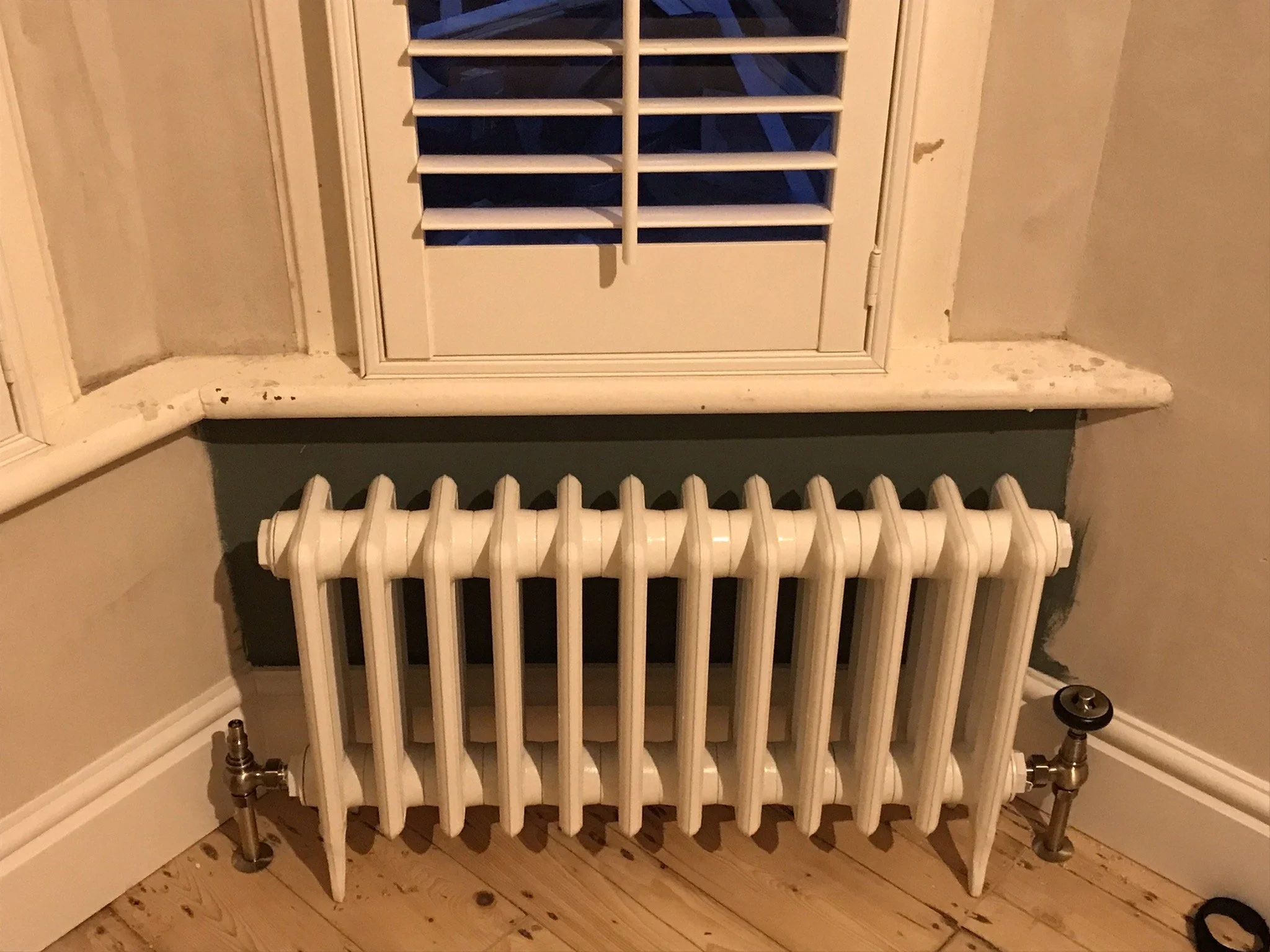A white radiator with multiple vertical columns is installed beneath a window with white shutters. The wall behind the radiator is painted gray, and the window has a white frame. The floor is wood, and there is white molding along the base of the wall.