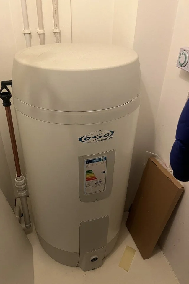 A tall, white, cylindrical hot water heater in a utility room, with a BP tag on the front and connected pipes on the side.