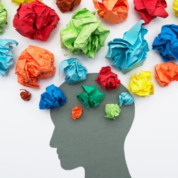 A paper cut-out of a human head silhouette with colorful crumpled paper balls inside and around it, representing thoughts or ideas.