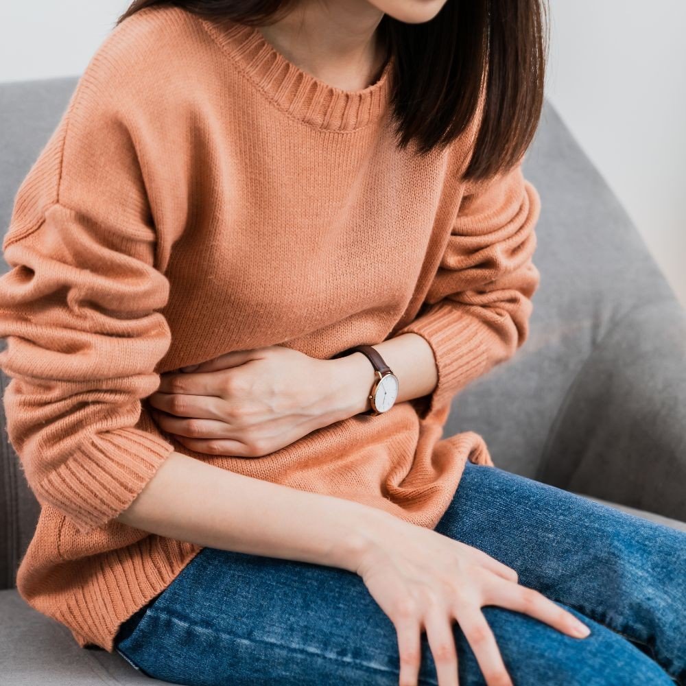 A woman sitting on a gray couch, wearing a peach-colored sweater and blue jeans. She has a wristwatch on her left wrist and is clutching her stomach with her right hand, indicating she may be experiencing stomach pain.
