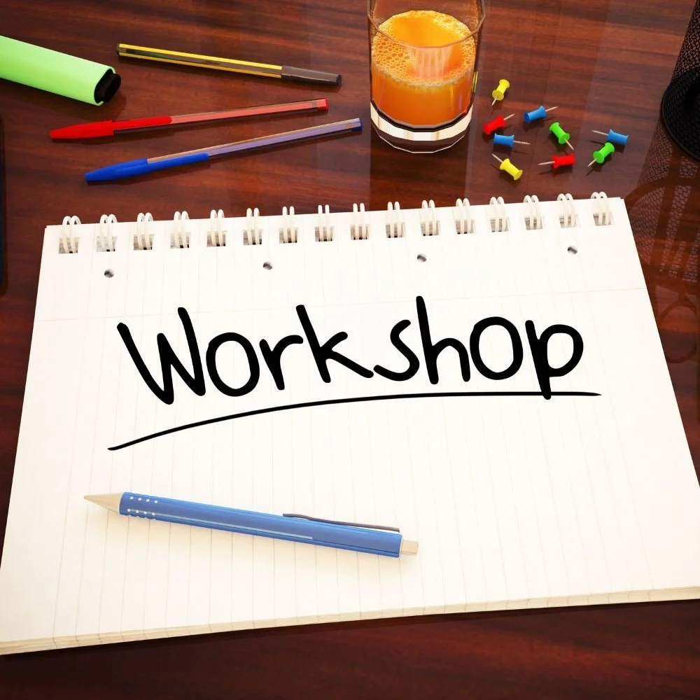 A spiral-bound notepad with the word 'Workshop' written on it, placed on a wooden desk along with a blue pen, a glass of orange juice, colorful push pins, and various pens and markers.