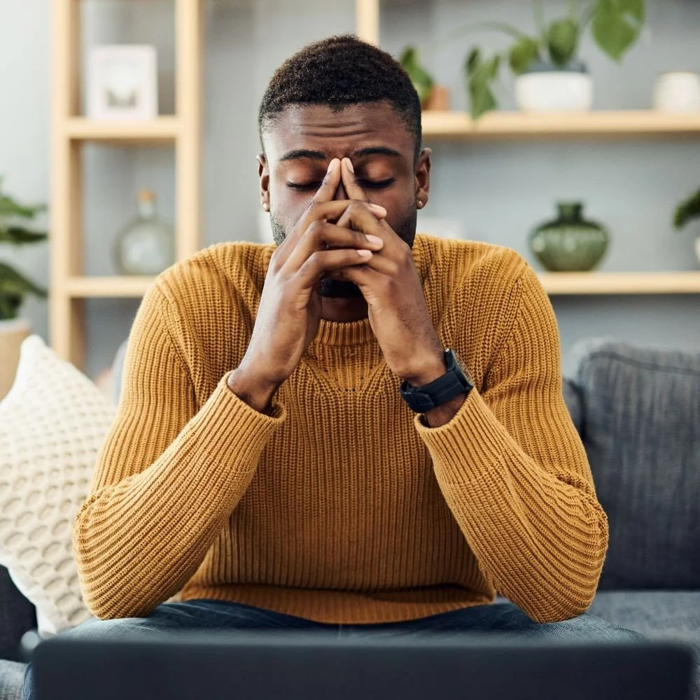 A man sitting on a couch with his eyes closed, holding his fingers together in front of his face as if in prayer or deep thought.