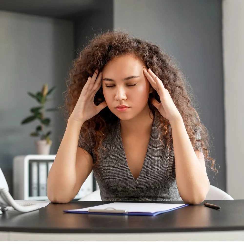 A young woman with curly hair is sitting at a desk with her eyes closed and hands pressed to her temples, appearing stressed or overwhelmed.