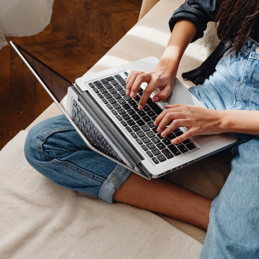 Person sitting on a couch typing on a laptop placed on their lap, wearing blue jeans and a black long-sleeve shirt.