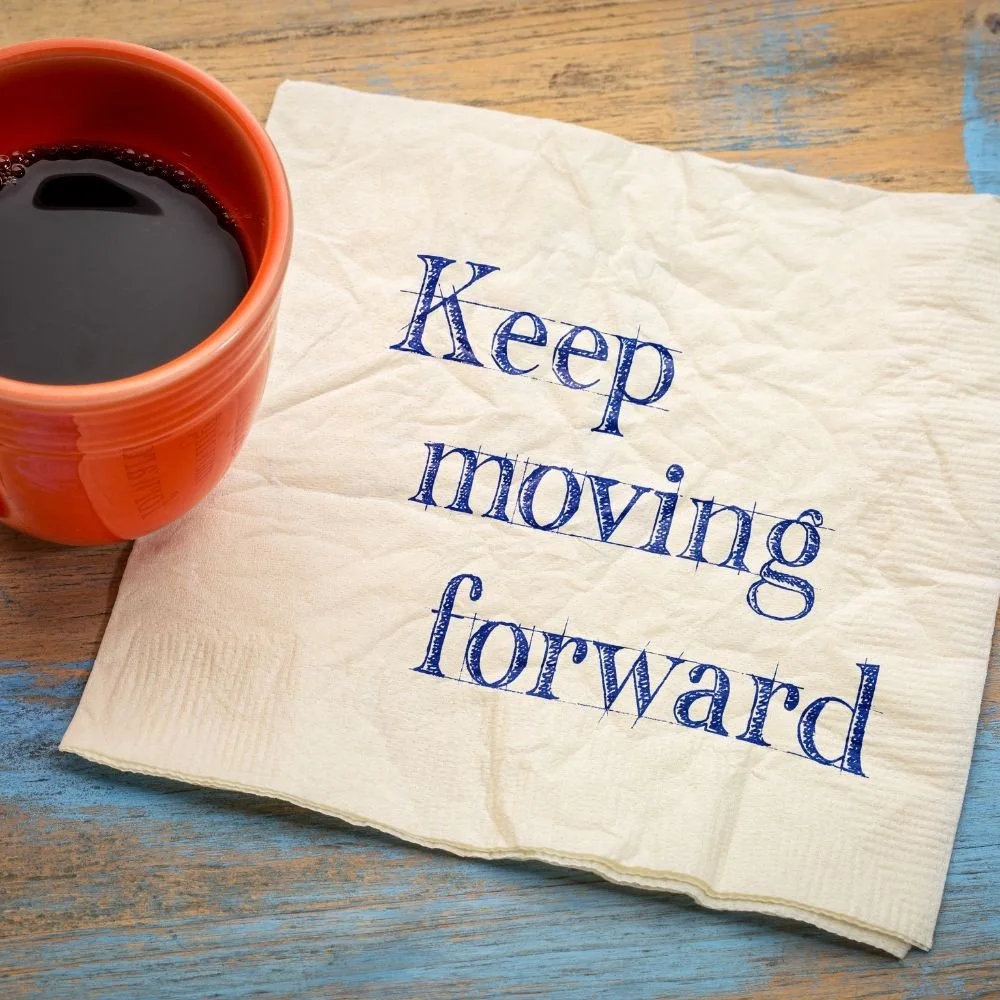 A cup of black coffee next to a crumpled napkin with the words "Keep moving forward" written in blue ink on a wooden table.