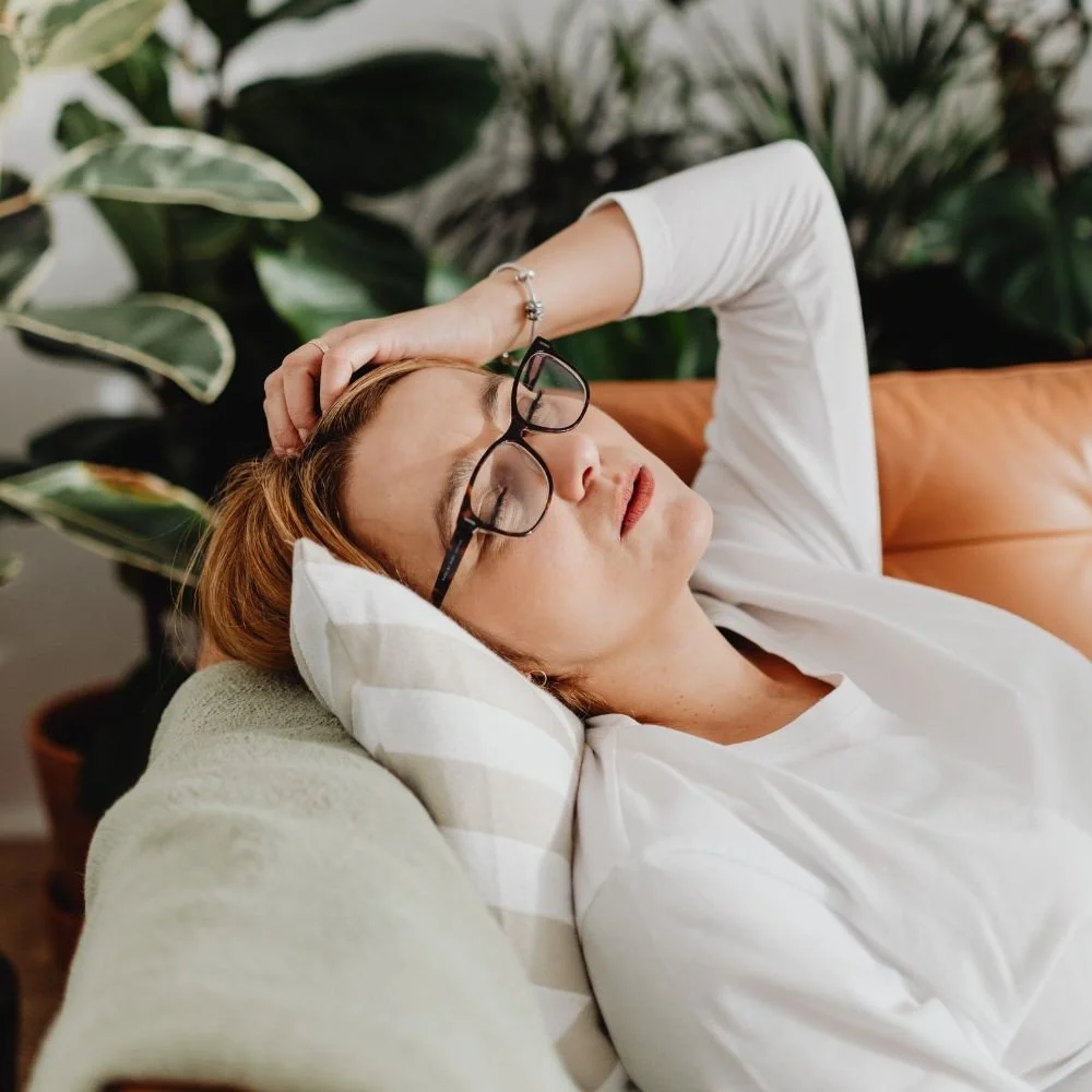A woman with glasses resting on a sofa with her eyes closed, head tilted back, and one hand on her head, in a cozy room with plants in the background.
