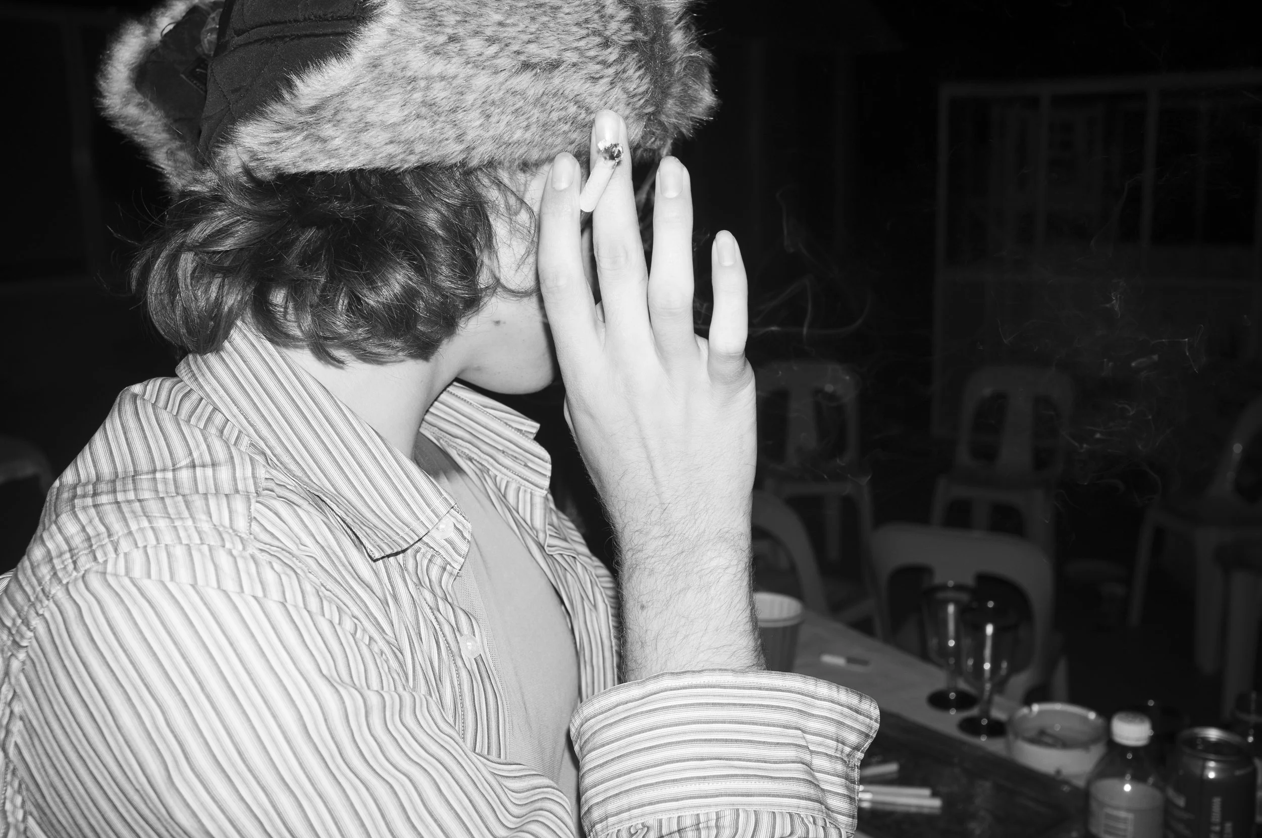 A person wearing a winter hat with fur trim and a striped shirt, seen in profile, is smoking a cigarette at a bar or restaurant.