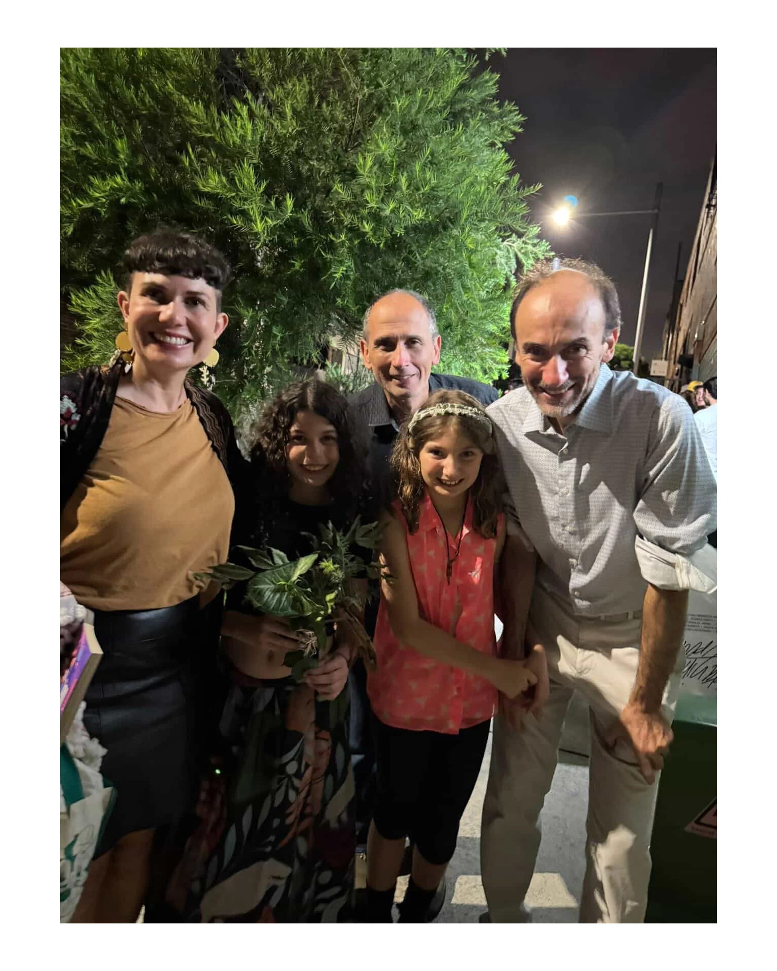 A group of people smiling and holding hands outdoors at night, with greenery and a streetlamp in the background.