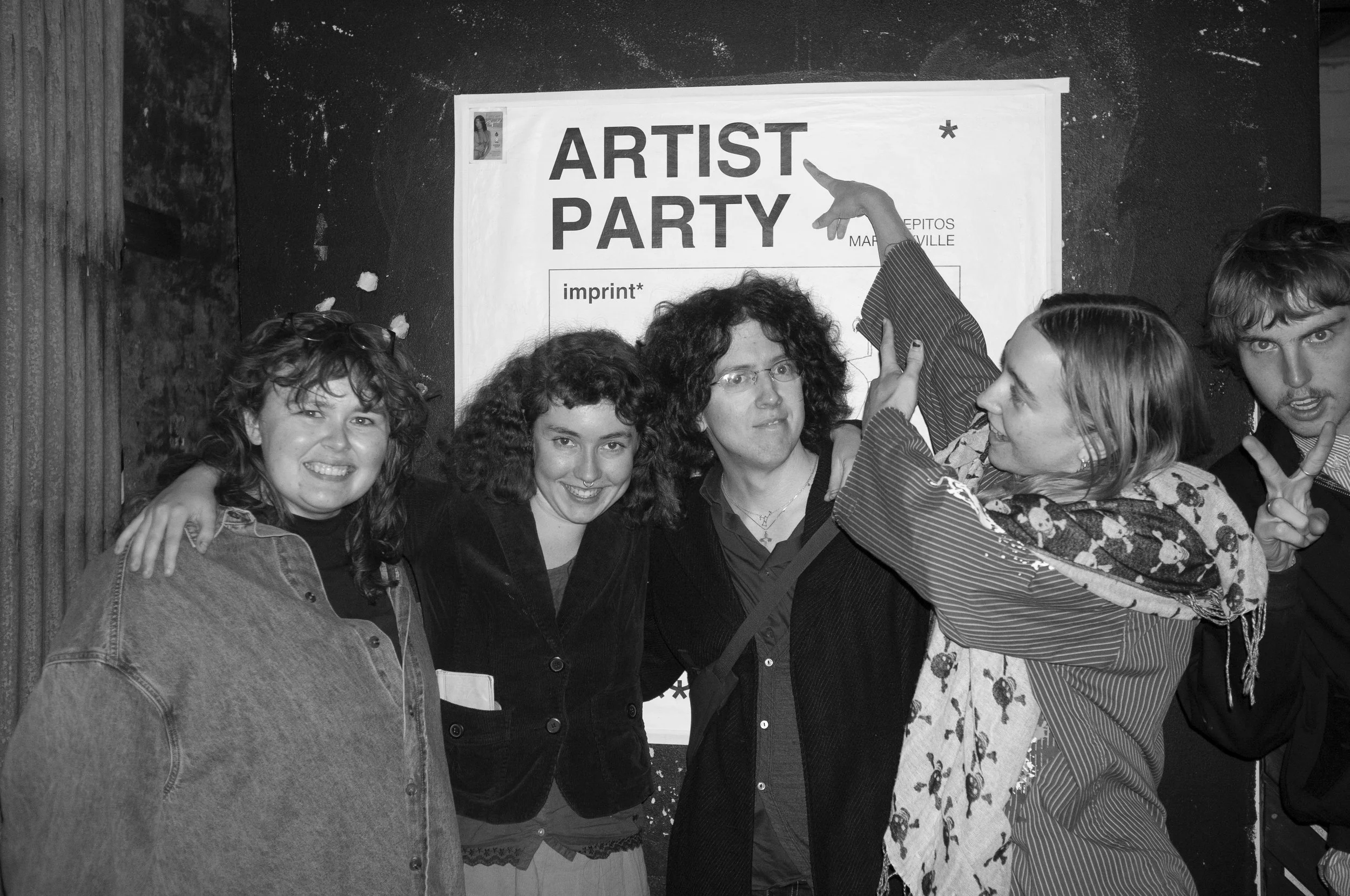 A group of six people posing in front of a sign that reads 'ARTIST PARTY.' They are smiling, and some are making gestures, standing close together against a dark wall.