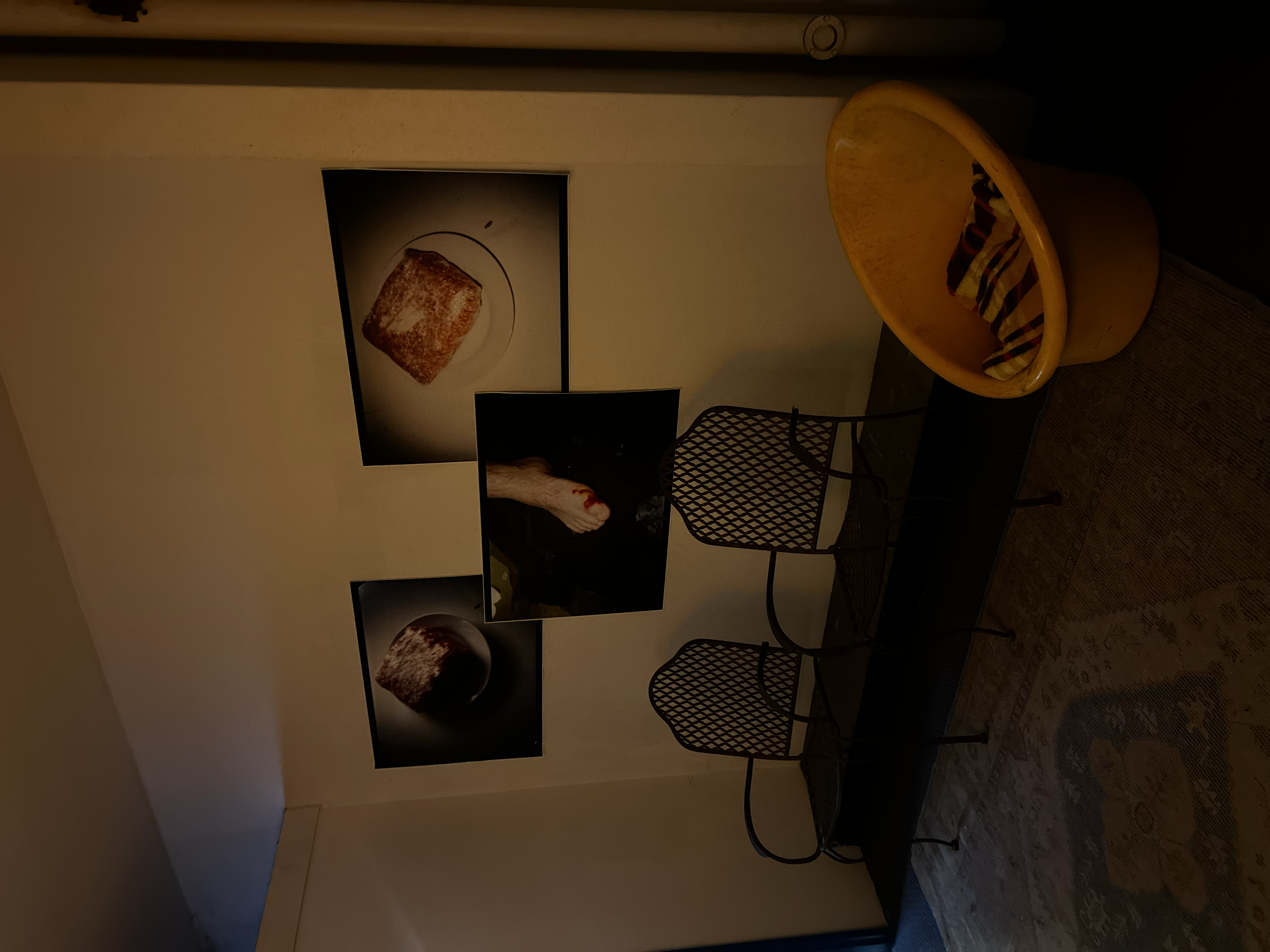 Interior room with a yellow cushioned chair, two metal chairs, a small table, and three framed photographs of food on the wall. One photo shows a piece of cake, another a bread loaf, and the third a bowl of food.