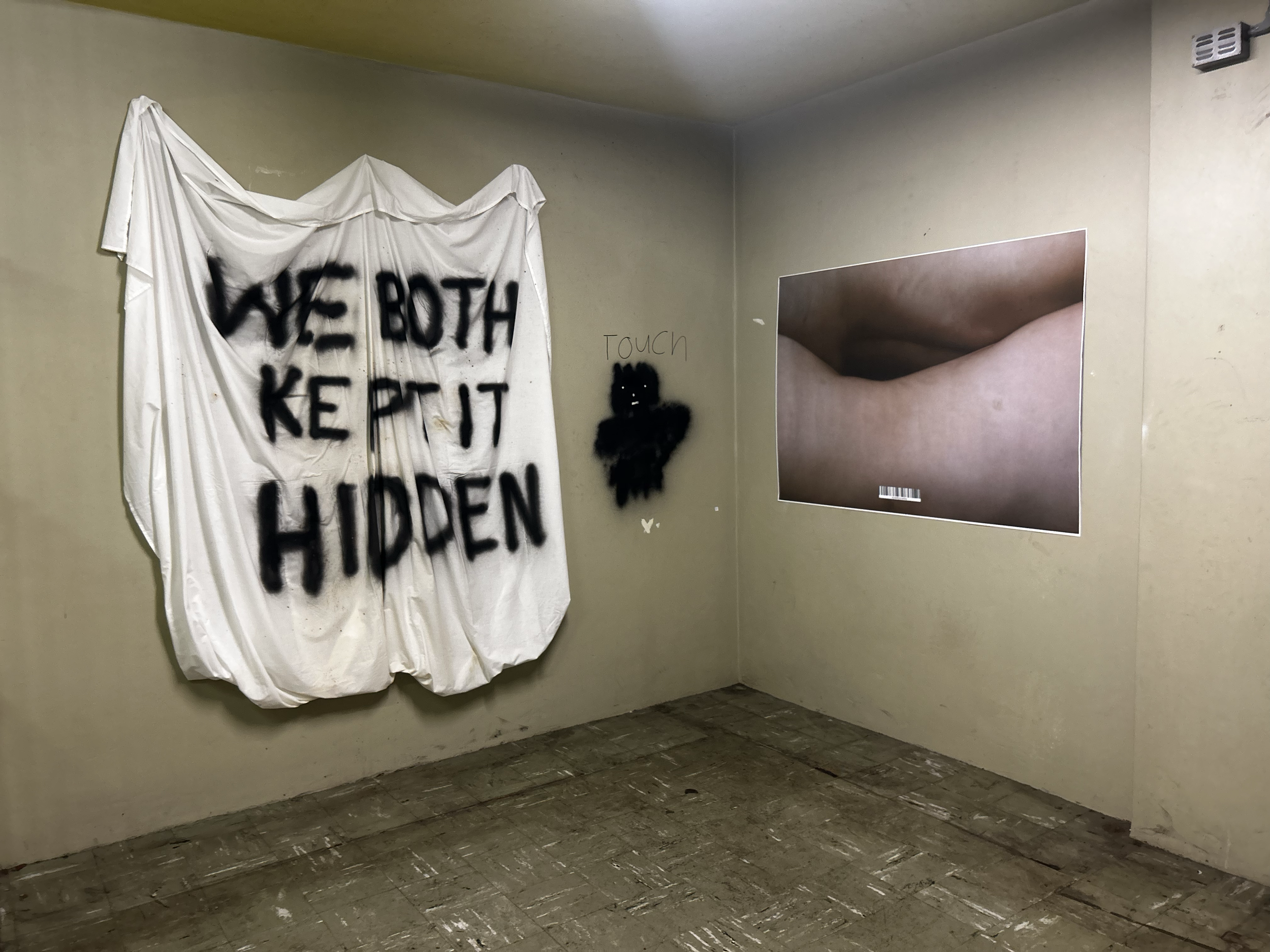 An art installation with a large white sheet draped on the wall with black spray paint reading "WE BOTH KEEP IT HIDDEN," and a smaller black spray-painted figure with the word "touch" written next to it. A photograph of a rolled-up dust on a surface 