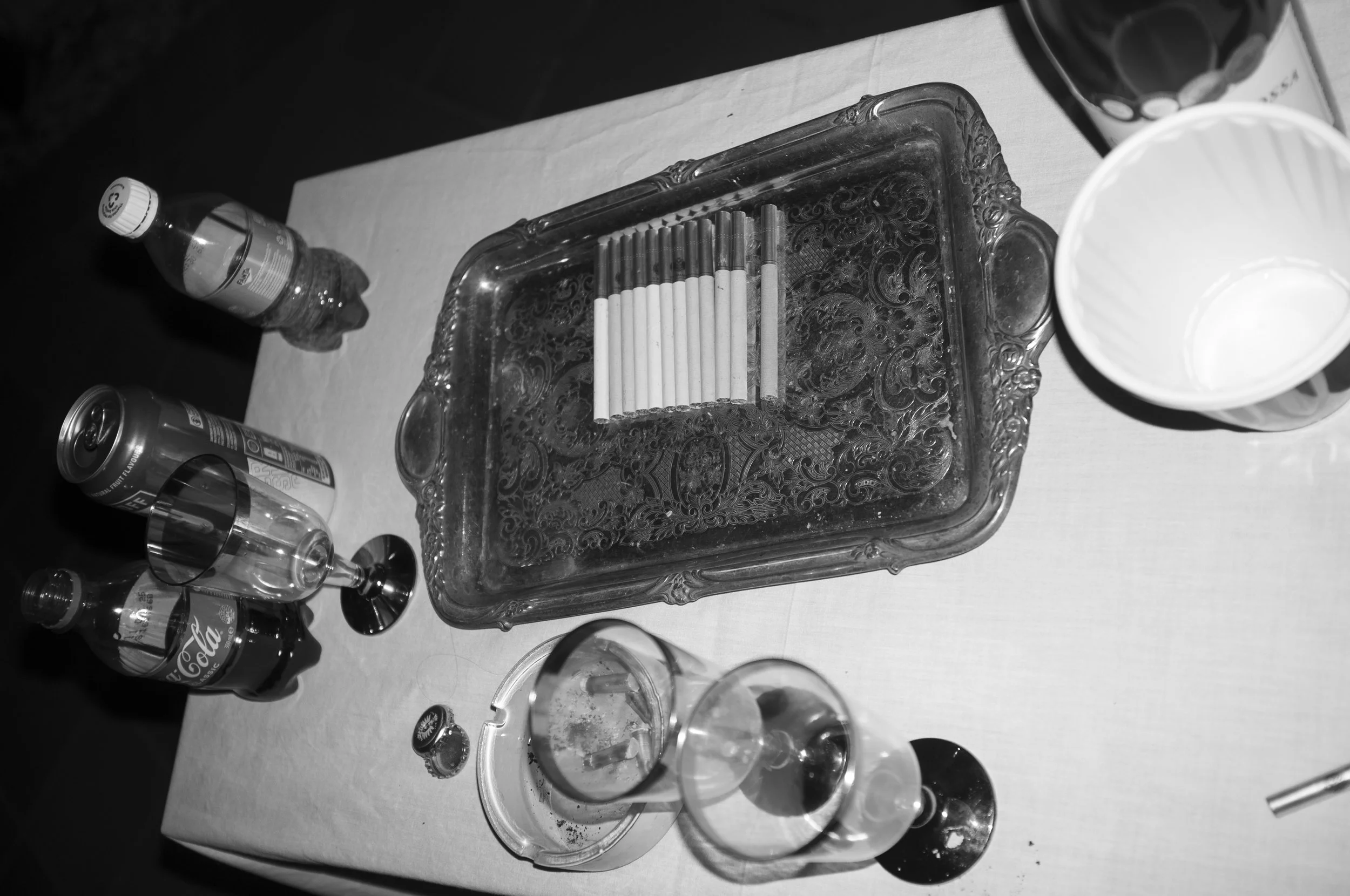 A table with a decorative tray holding cigarettes, an empty cup, a bottle of soda, a Coke can, some glasses, and an open bottle of water.
