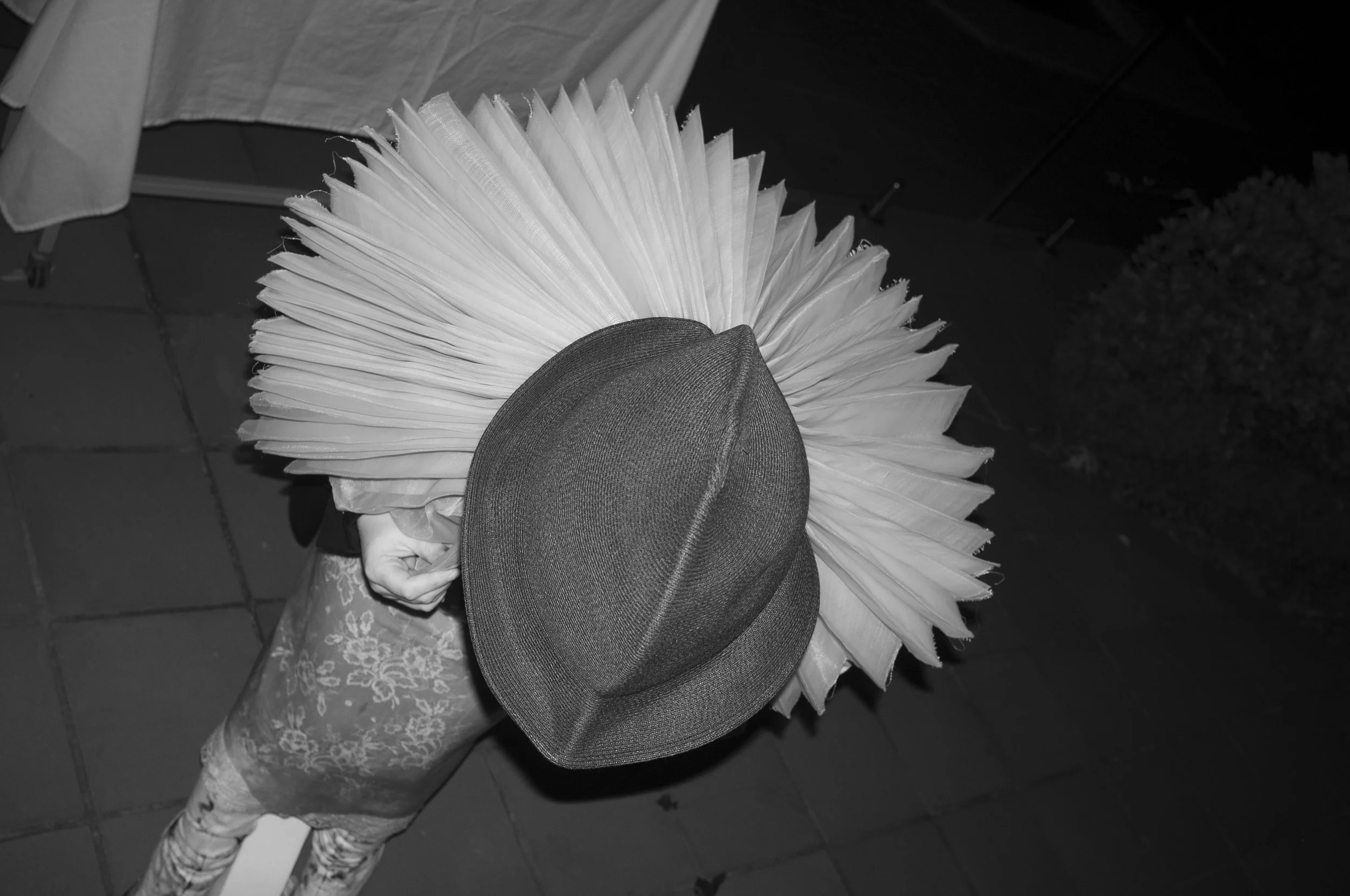 A person wearing a large pleated skirt, floral-patterned tights, and a hat, standing on a tiled floor, with a table and bushes visible in the background.