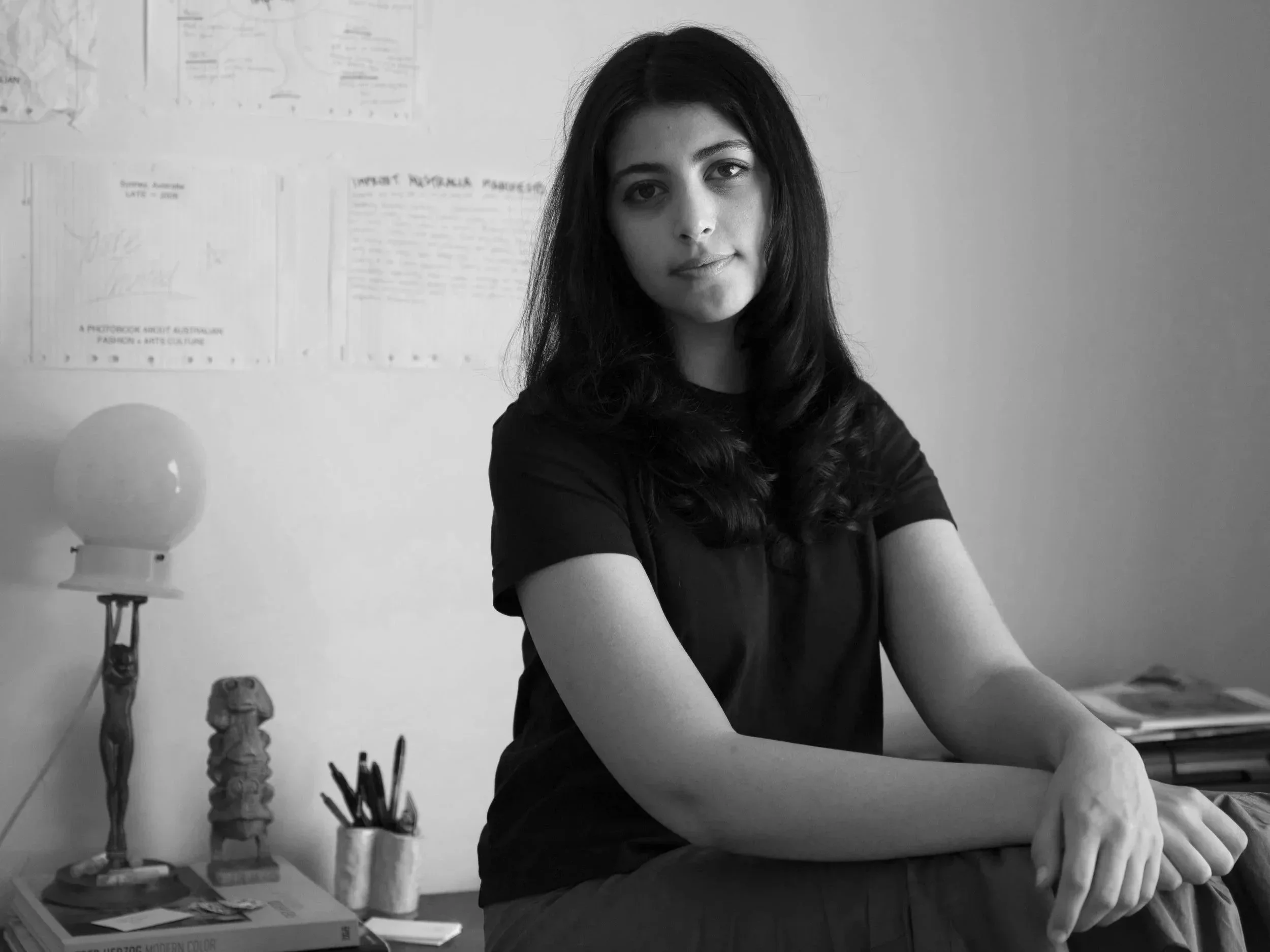 A young woman with long dark hair sits on a desk in a room with papers and drawings on the wall and objects on the desk, dressed in a black t-shirt.