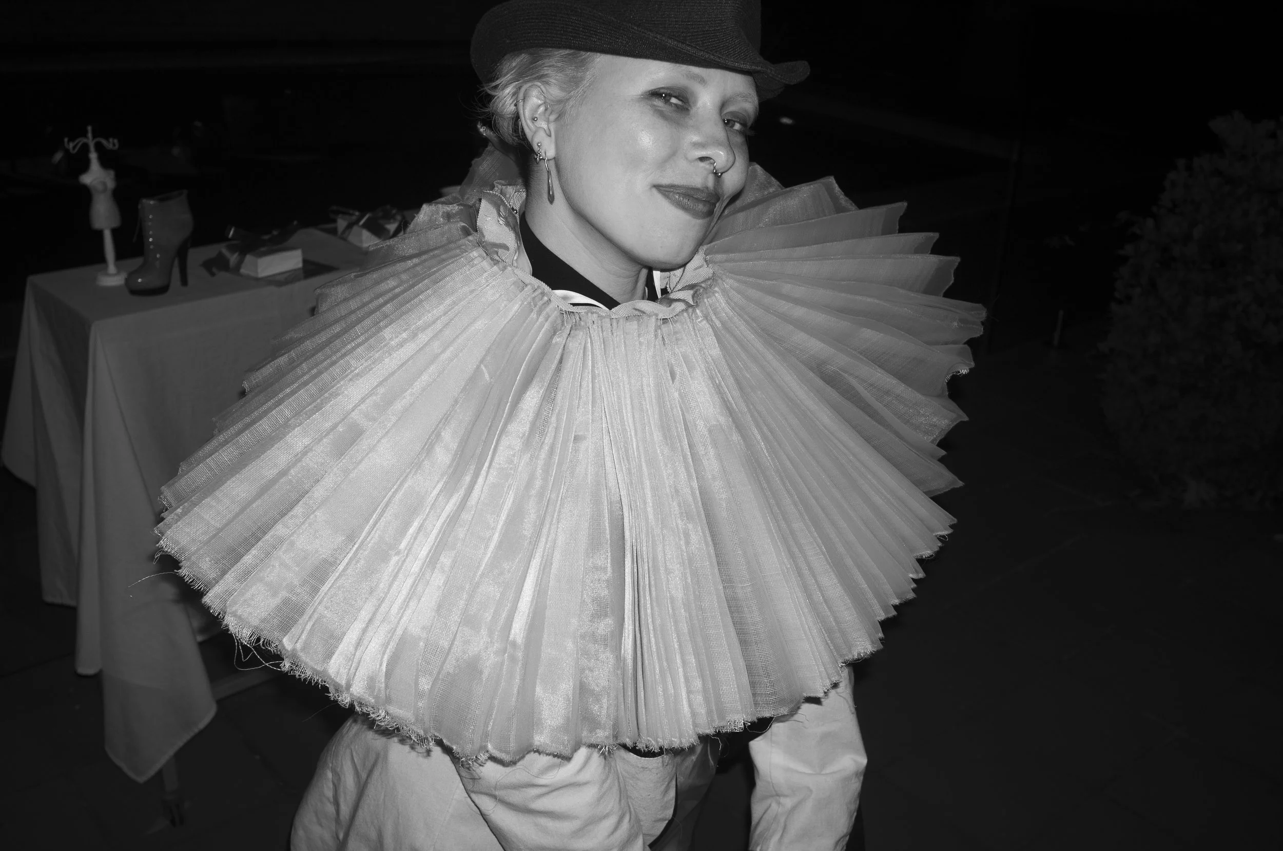 A woman wearing a large, ruffled, pleated collar costume, smiling, at an event with displayed shoes and accessories on a table in the background.