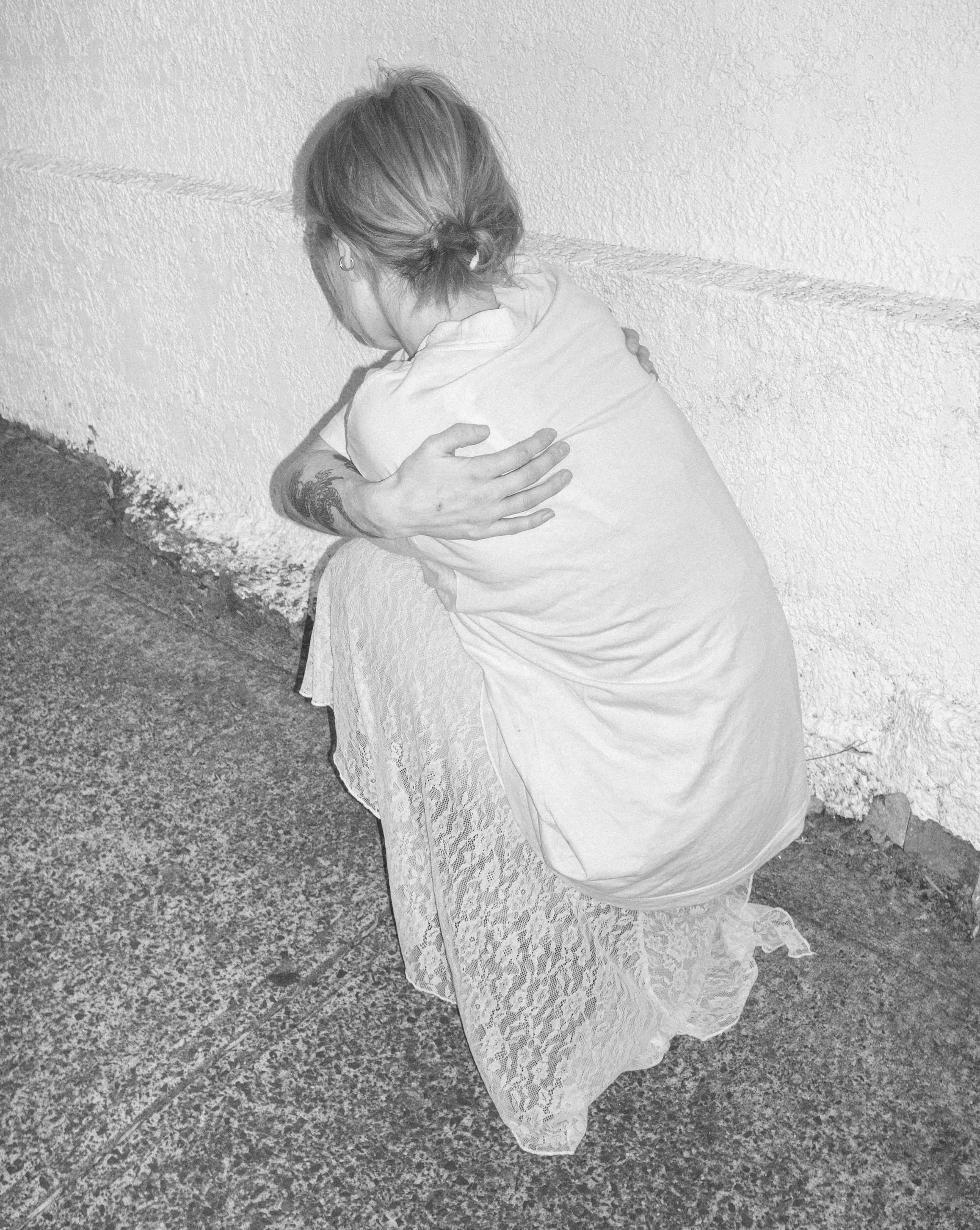 A woman in a white dress sitting on the ground against a wall with her arms crossed over her knees, looking downward.