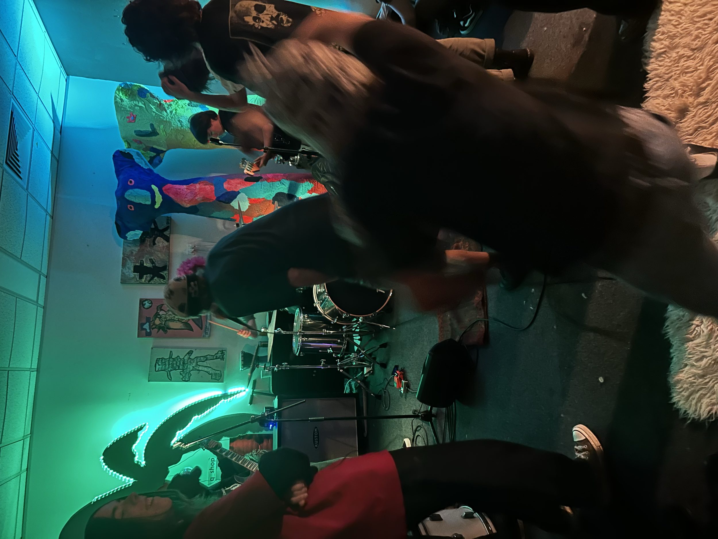 People playing musical instruments and singing in a dimly lit room decorated with colorful art on the walls and neon lights, creating a lively and artistic atmosphere.
