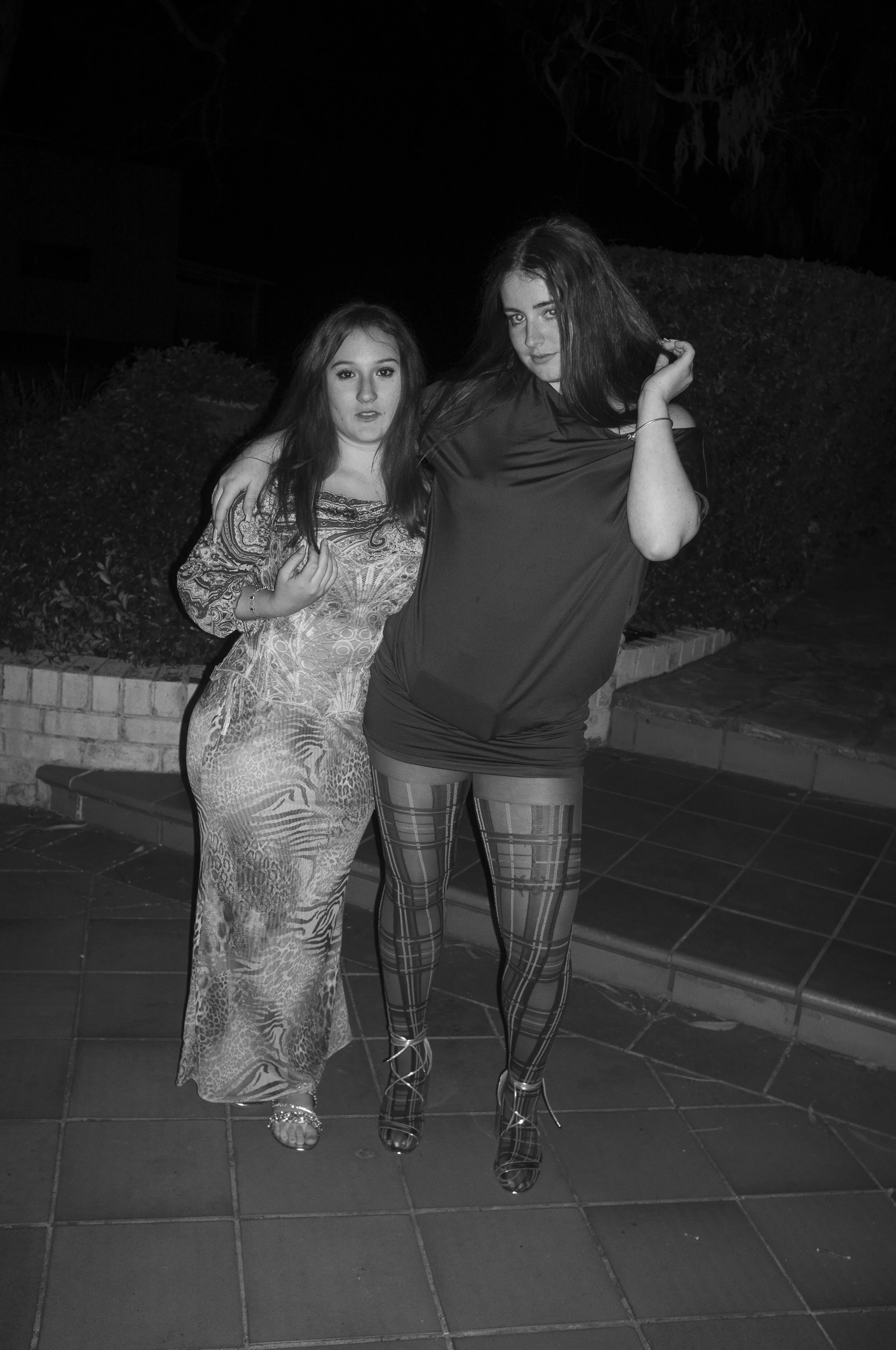 Two women are posing together outdoors at night, one wearing a patterned dress and the other wearing a short dress with patterned tights, standing on a tiled area near some steps and bushes.