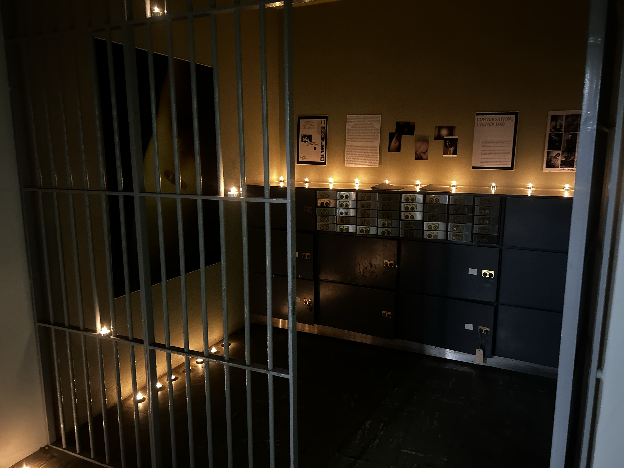 A dark room with a metal grate door in the foreground leading into a space decorated with small candles, posters, and switches or electrical panels on the wall.