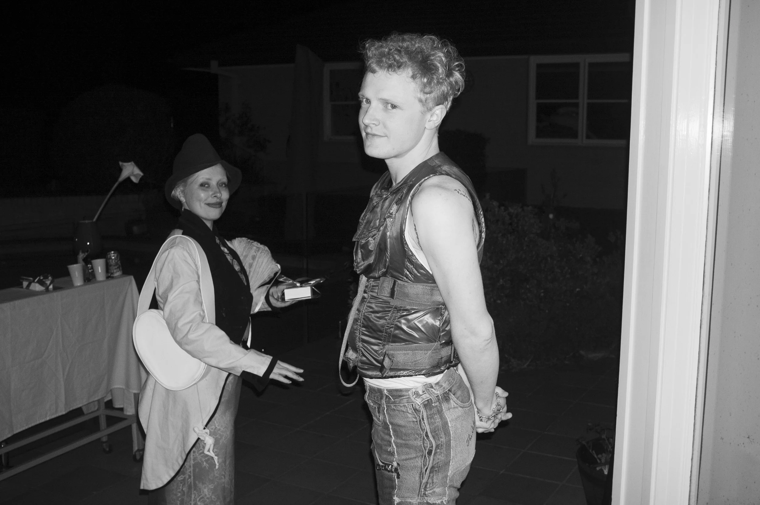 A young man with short, curly hair standing with his hands behind his back, wearing a shiny sleeveless top and ripped jeans, looking at the camera, with a woman dressed as a witch in the background, smiling, wearing a witch's hat, costume, and makeup